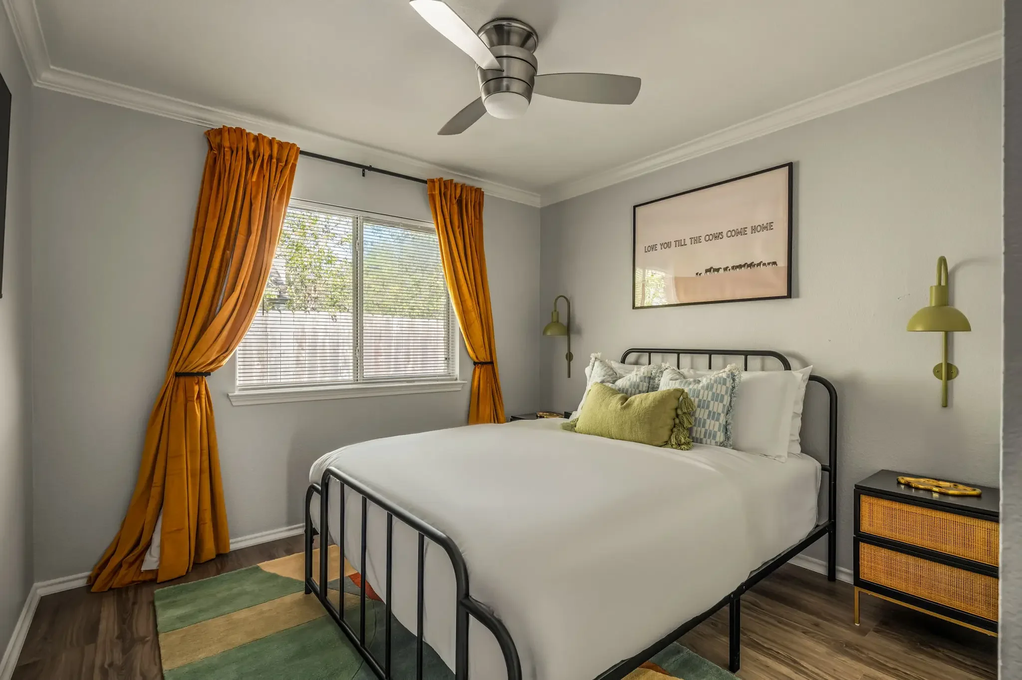 Short-term rental bedroom design with gold curtains, black iron bed frame, green pillows, and rattan nightstand — Austin TX.