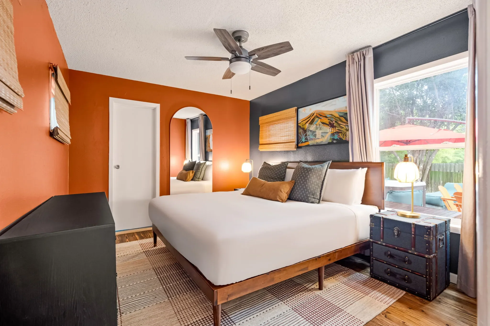 Airbnb bedroom design with orange and navy accent walls, wood bed frame, trunk nightstand, arched mirror, and backyard access — Austin TX.