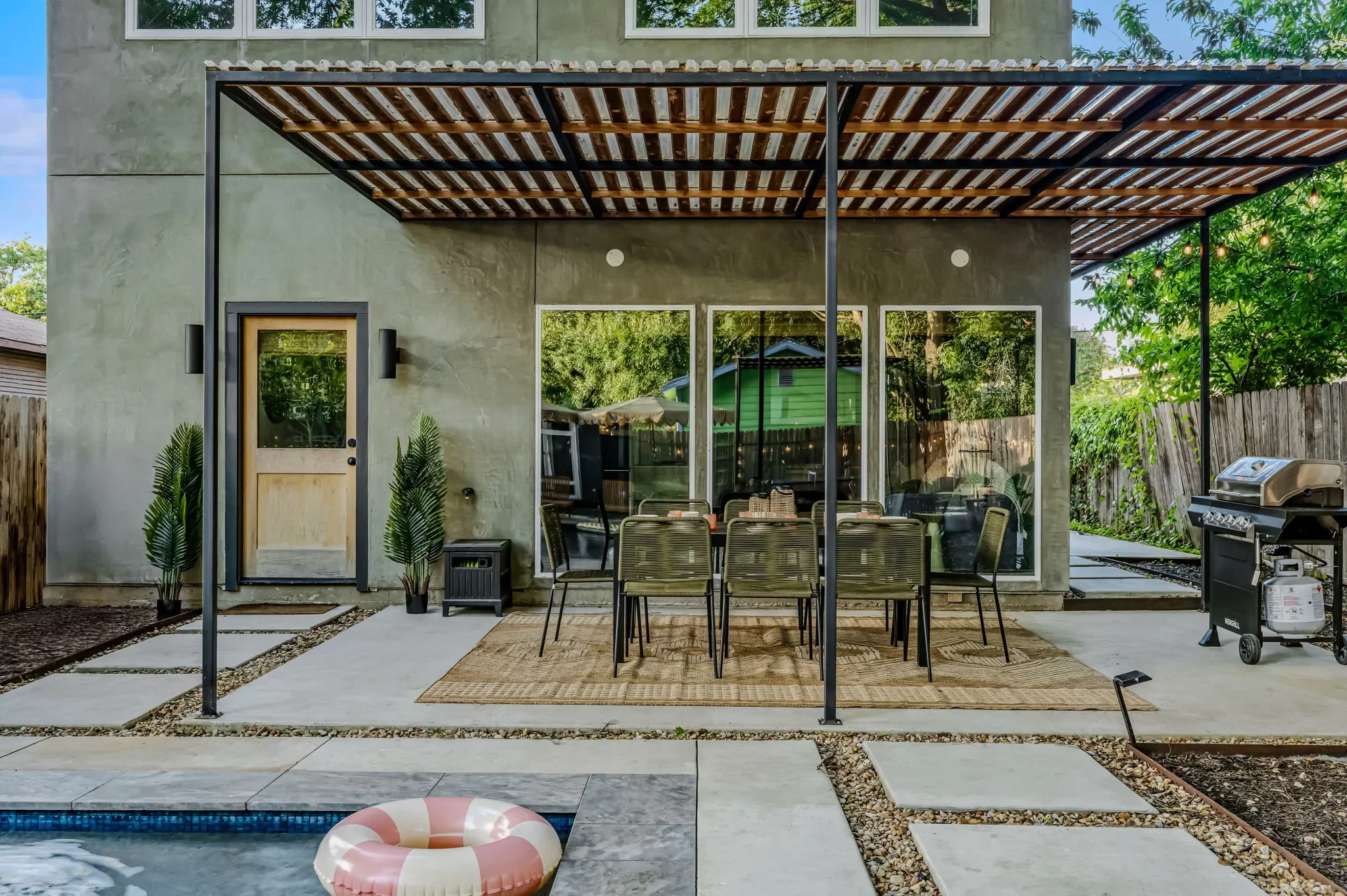 Short-term rental outdoor dining design with steel pergola, long dining table, rattan chairs, and grill station beside pool — Austin TX.