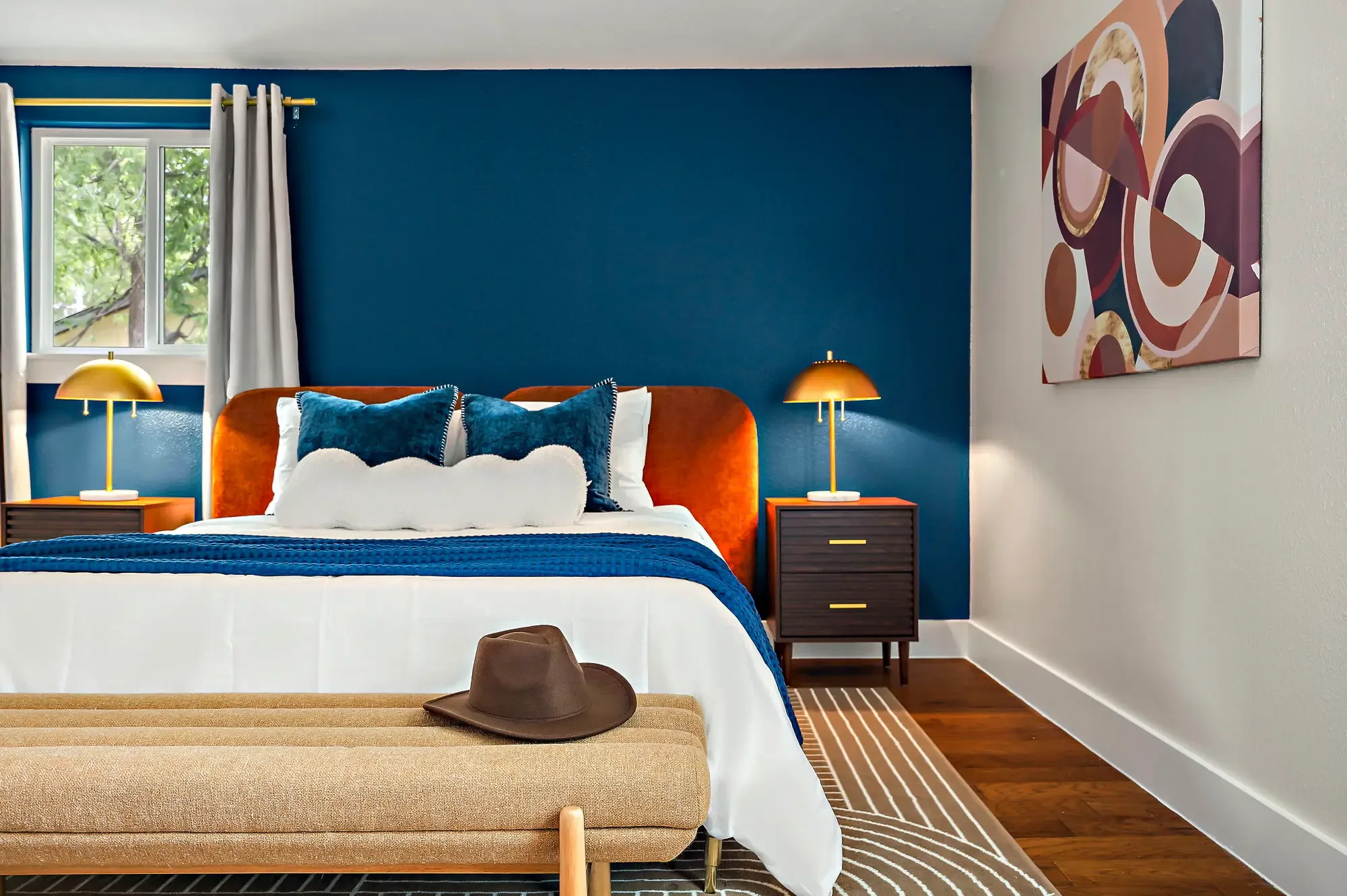 Short-term rental bedroom design with navy accent wall, orange velvet headboard, gold lamps, blue pillows, and cowboy hat styling — Austin TX.