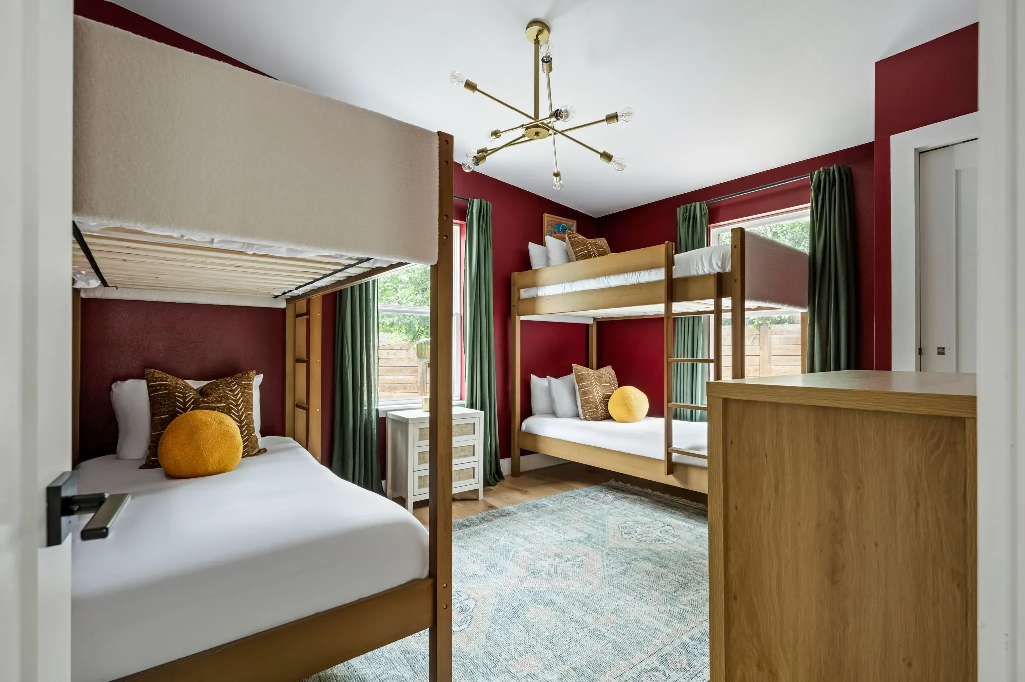 Short-term rental bunk room design with bold red walls, wood bunk beds, green curtains, and brass sputnik chandelier — Austin TX.