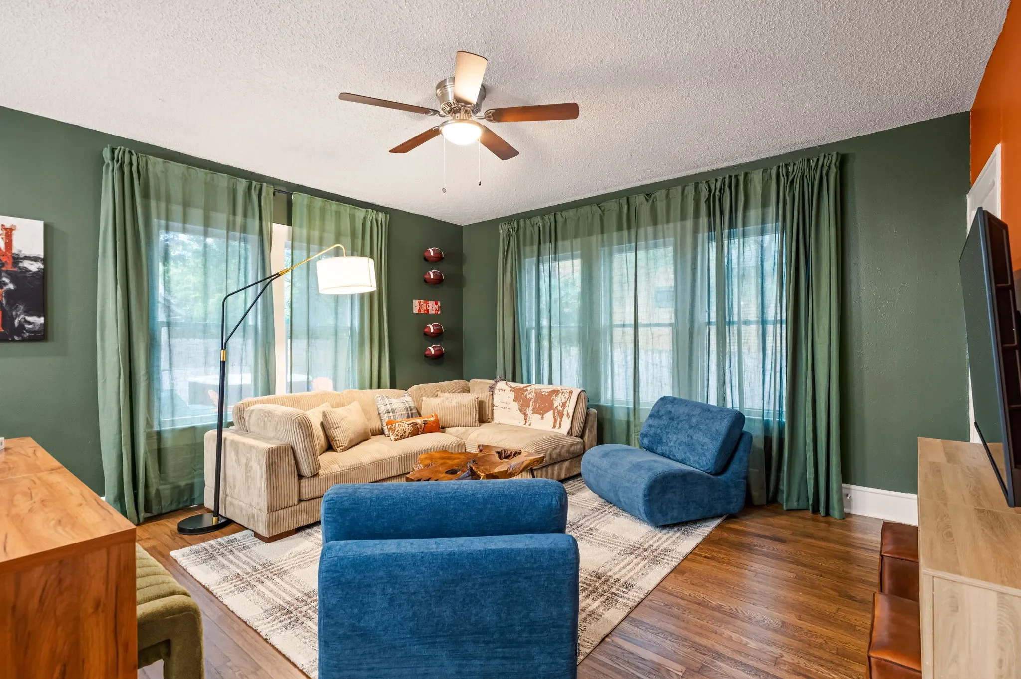 Airbnb living room design with dark green walls, tan corduroy sectional, blue accent chair, football wall decor, and green curtains — Austin TX.