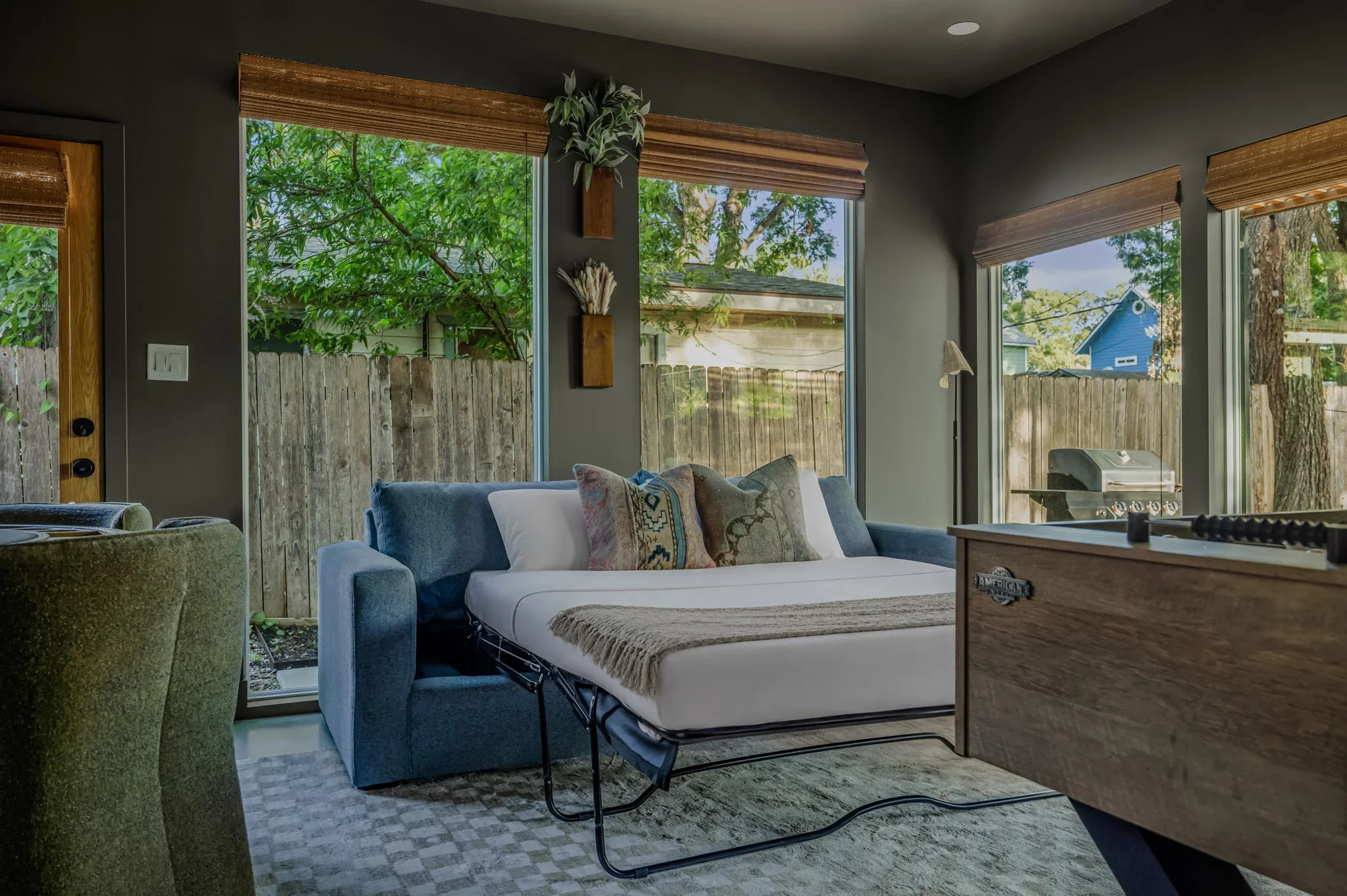 Short-term rental game room design with sleeper sofa pulled out as extra bed, olive green walls, and garden view — Austin TX.