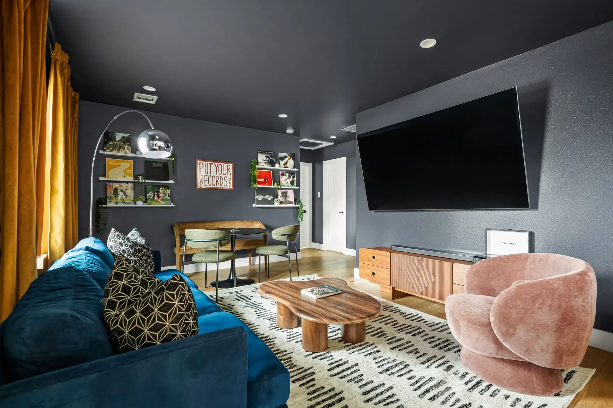 Short-term rental lounge room design with dark walls, blue velvet sofa, large TV, vinyl record shelves, and gold curtains — Austin TX.
