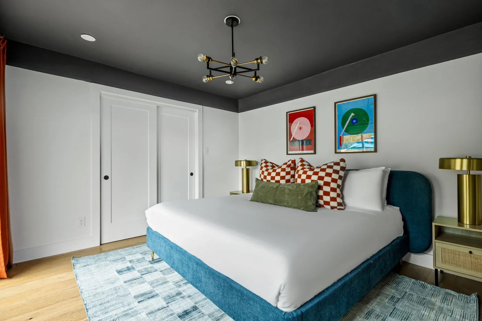 Short-term rental guest house bedroom design with dark ceiling, blue upholstered bed, checkered pillows, brass lamps, and colorful wall art — Austin TX.