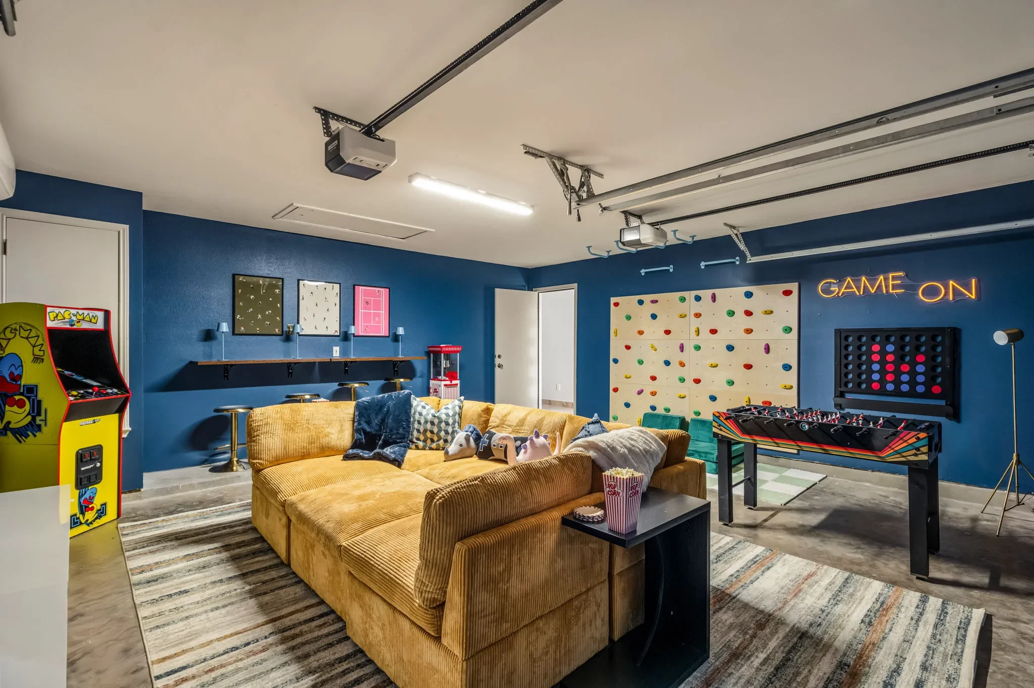 Converted garage game room with blue painted walls, arcade games, game tables, a giant yellow sectional, and a neon “Game On” sign.