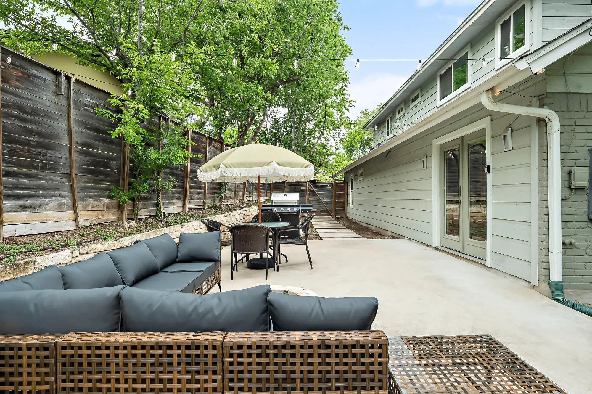 Airbnb backyard design with dark sectional, macrame umbrella, dining area, grill, and string lights under mature trees — Austin TX.