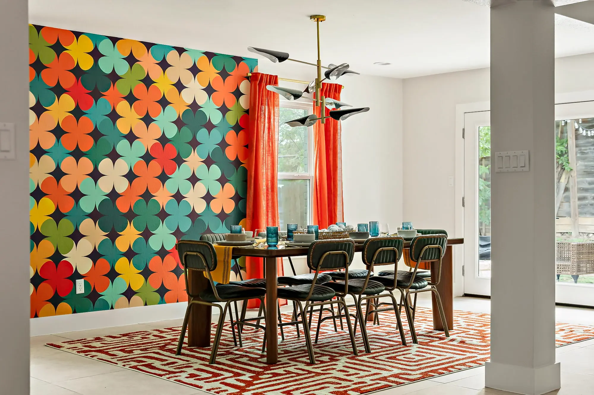 Mid-century modern dining room with geometric wallpaper, bright orange floor-to-ceiling curtains, red leather area rug, and a large dining table for hosting.