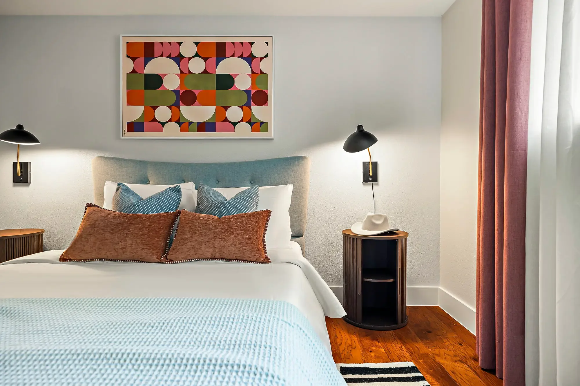 Short-term rental bedroom design with teal upholstered headboard, retro circle artwork, black sconces, and round nightstand — Austin TX.