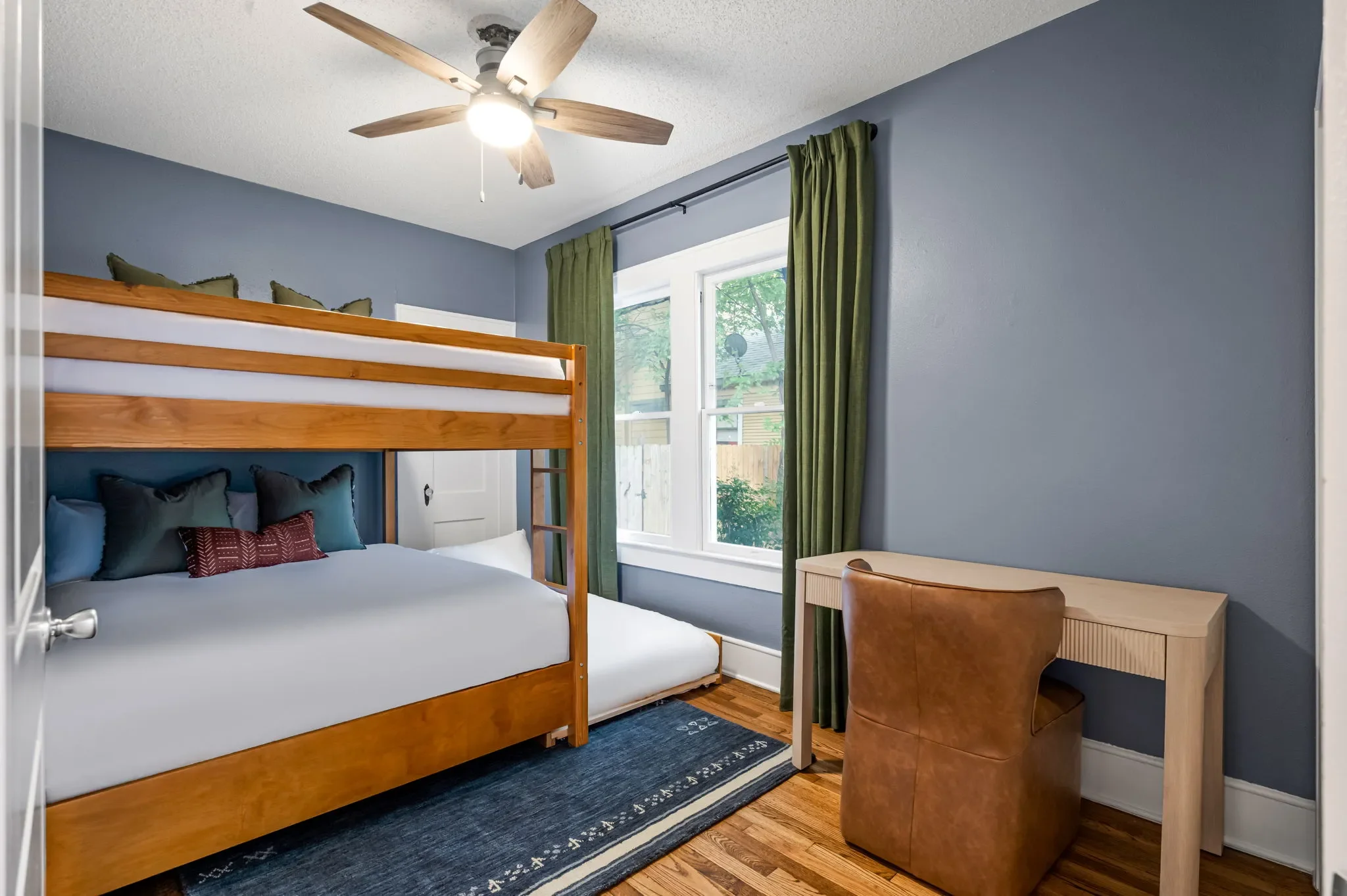 Short-term rental bunk room design with wood bunk beds, blue-gray walls, green curtains, leather bean bag, and framed game art — Austin TX.