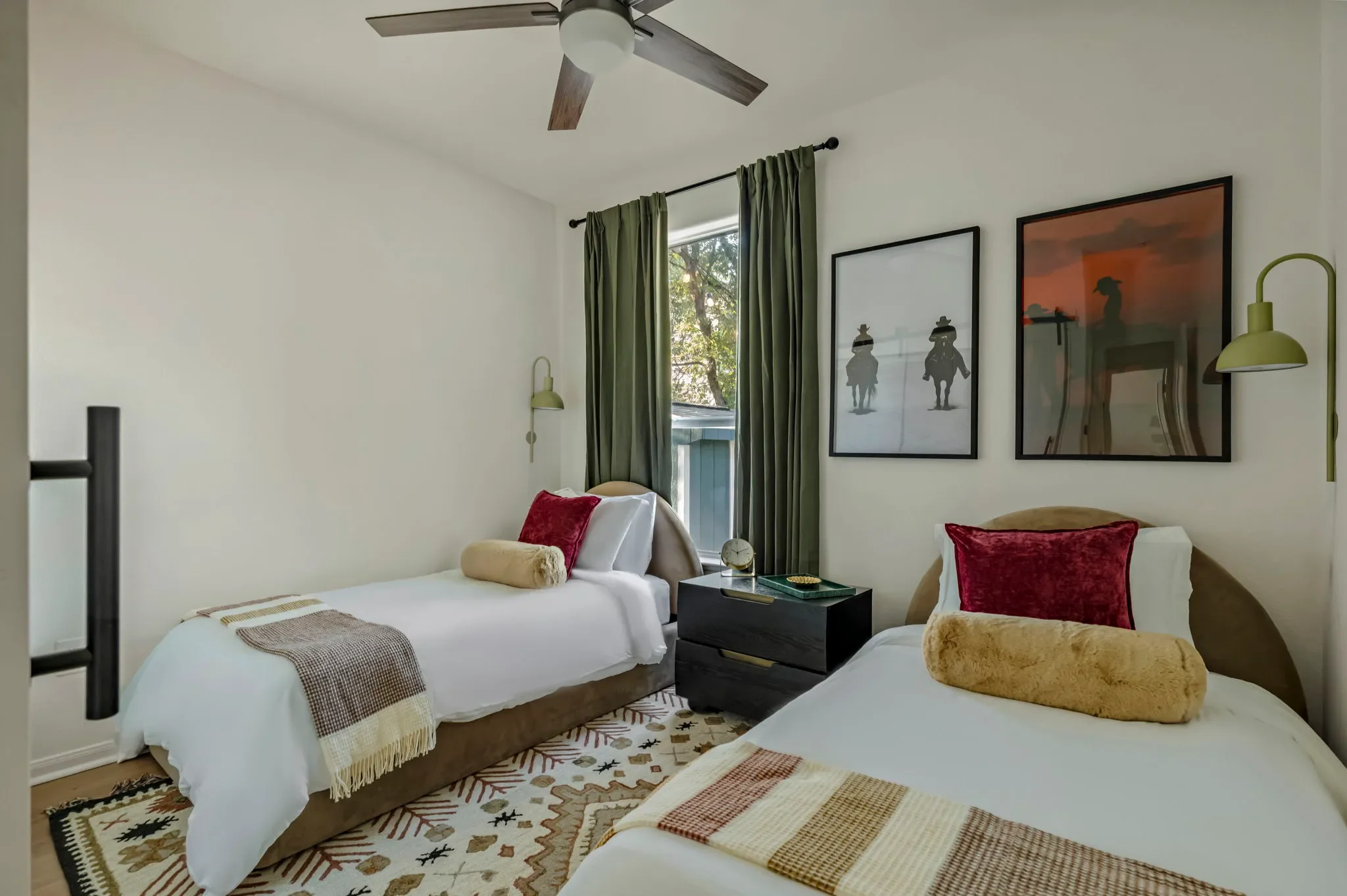 Bedroom with twin beds, cowboy western wall art, green wall sconces, boho western-style area rug, floor-to-ceiling olive green curtains, and a black nightstand.