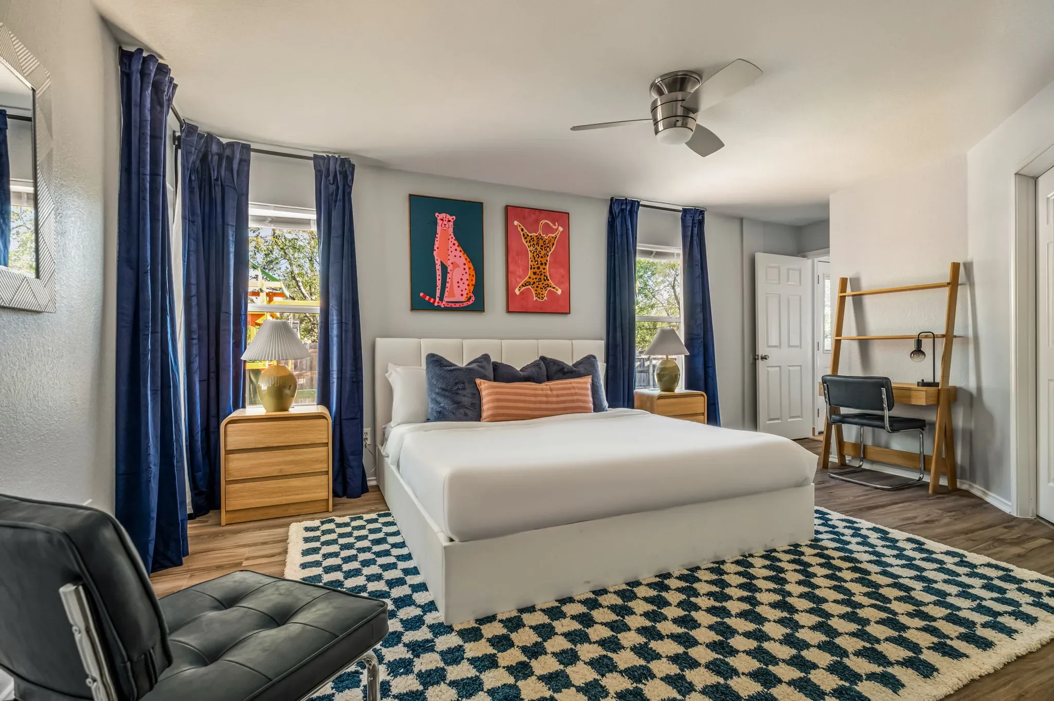 Bedroom with an off-white king bed, blue floor-to-ceiling curtains, arched wooden nightstands, animal wall art, and a blue checkered rug.