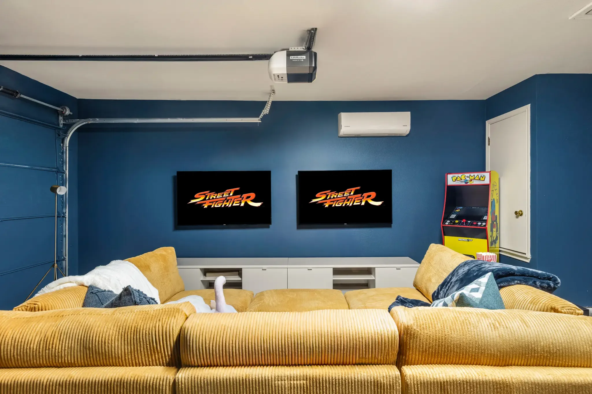Airbnb game room design with dual TVs, blue accent wall, yellow sectional, and gaming setup — Austin TX.