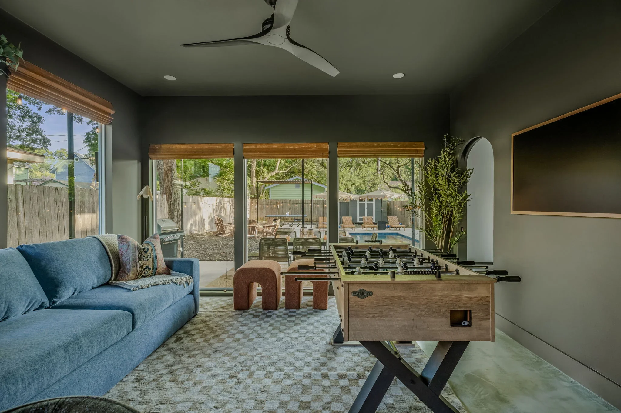 Airbnb game room design with deep olive green walls, blue sofa, foosball table, TV, and sliding glass doors to pool — Austin TX.