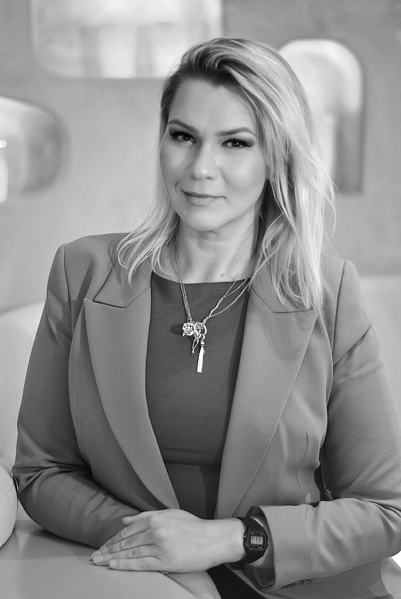 Black and white photo of a woman with shoulder-length hair, wearing a blazer and a necklace, seated at a table.