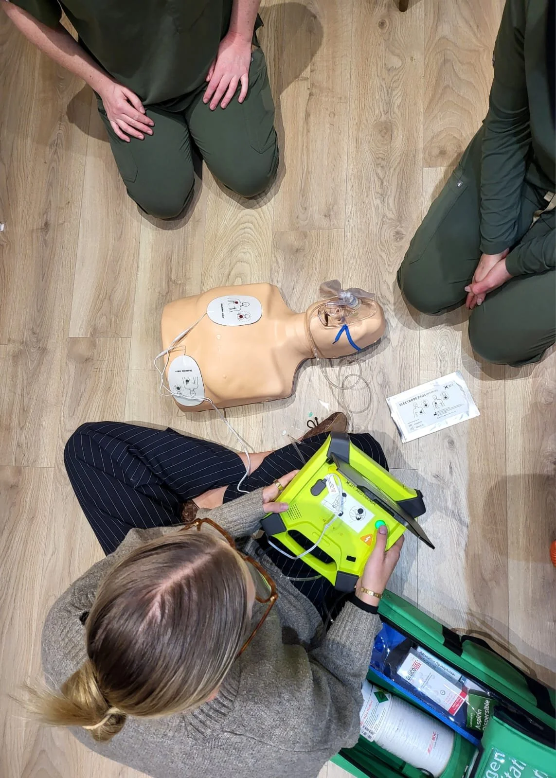 A woman demonstrating CPR using an emergency defibrillator on a training mannequin while two individuals kneel nearby observing.
