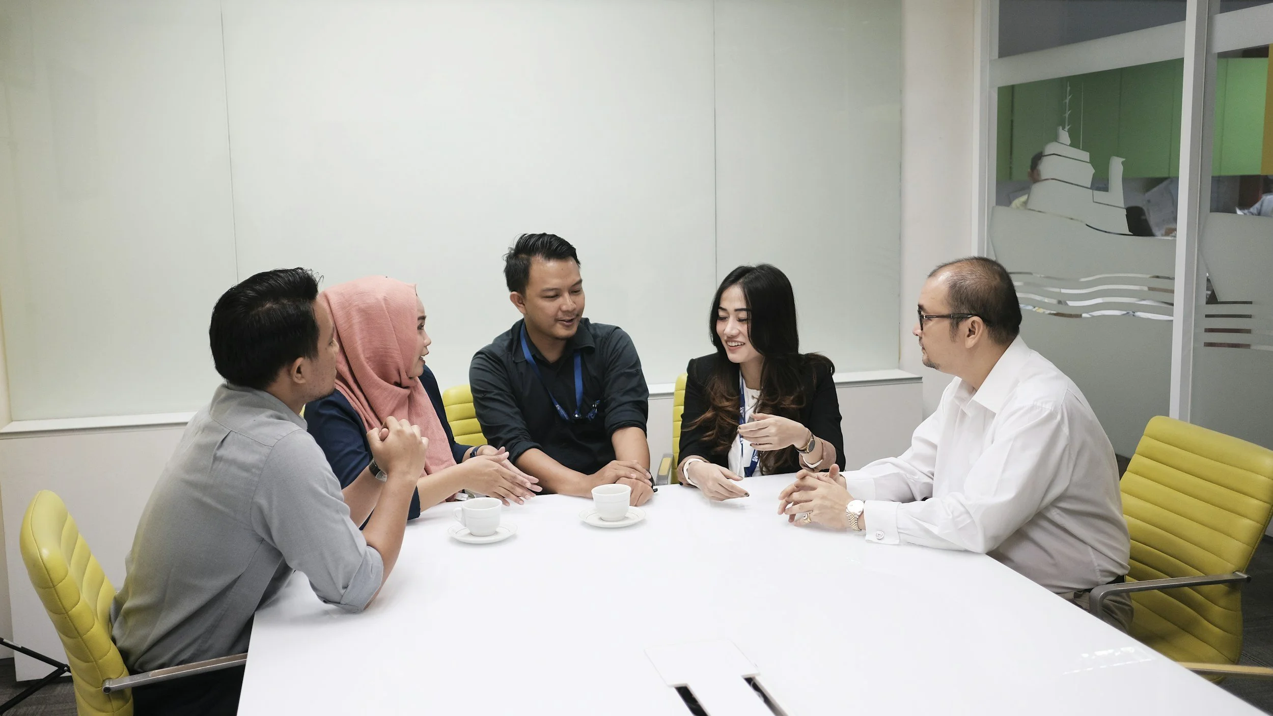 Five diverse professionals having a meeting in a modern office conference room, sitting around a white table with cups of coffee, engaged in a discussion and smiling.