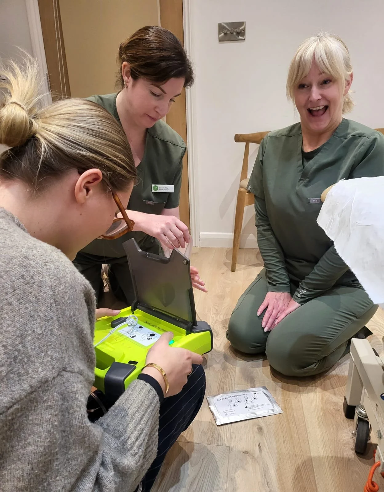 Three women in medical scrubs and one girl in a sweater, sitting on a wooden floor, practicing the use of an automated external defibrillator (AED).