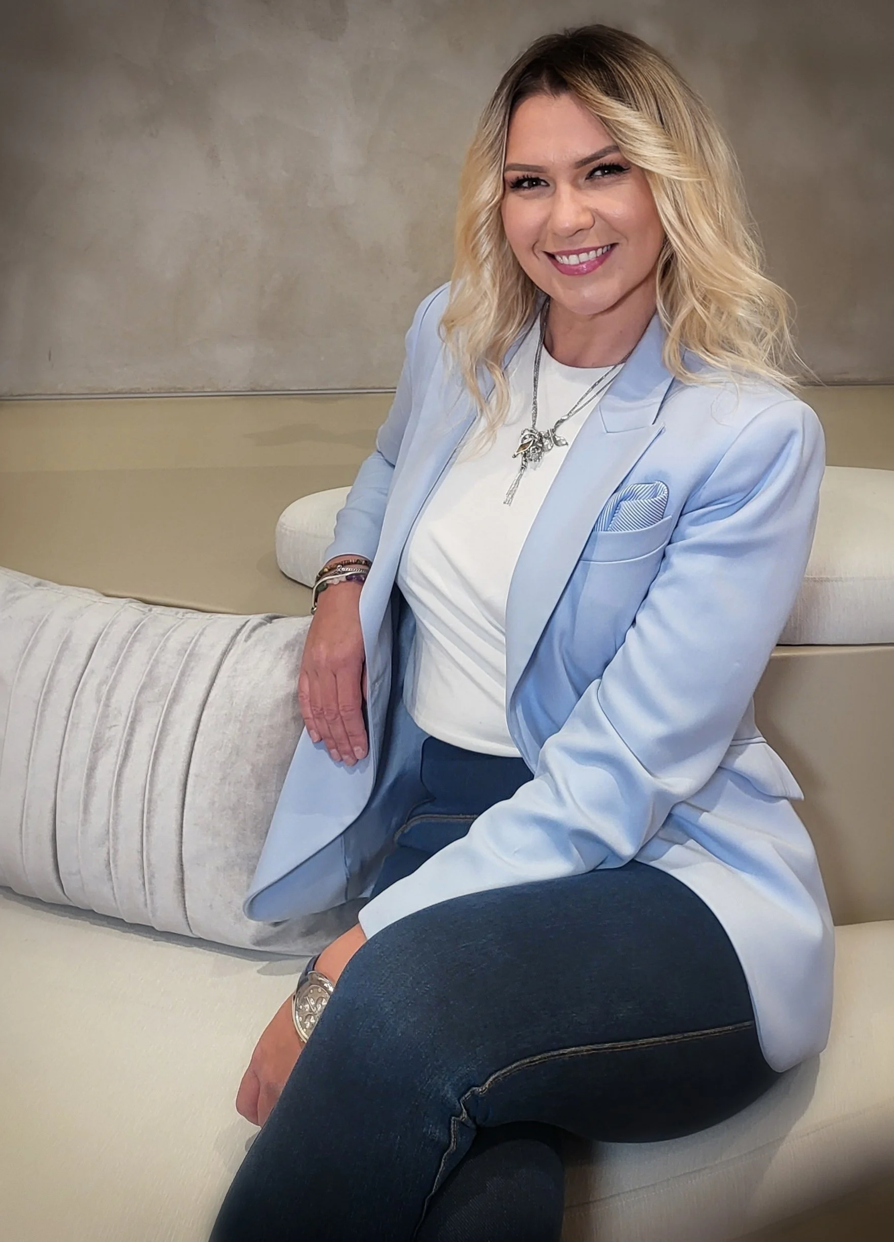 A woman with blonde hair and a bright smile, wearing a light blue blazer, white T-shirt, and dark jeans, sitting on a cream-colored sofa with a modern interior background.