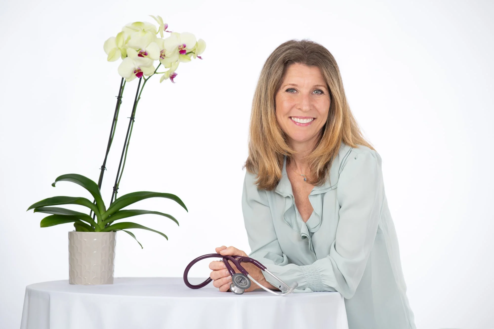 Dr. Jennifer Kostacos sitting at a round table with a white tablecloth, smiling, with a stethoscope in her hand, next to a potted orchid plant with white flowers and pink centers against a plain white background.
