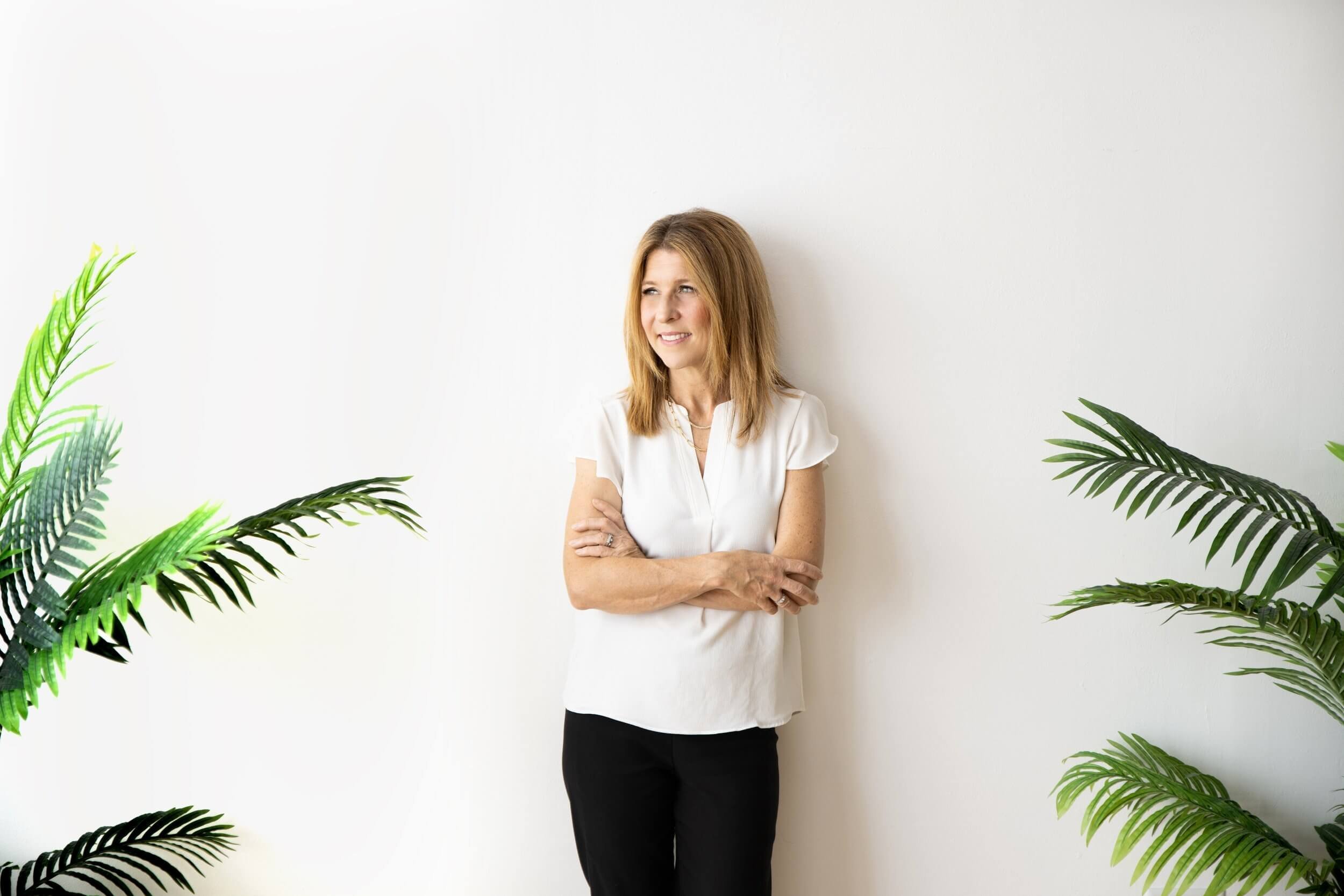Dr. Jennifer Kostacos wearing a white blouse and black pants, standing with arms crossed against a white wall, flanked by green potted plants.