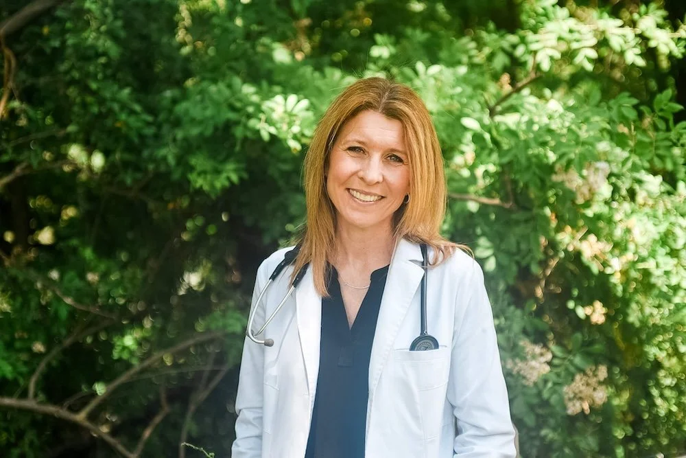 A smiling Dr. Jennifer Kostacos in a white coat with a stethoscope around her neck standing outdoors with green leafy trees in the background.