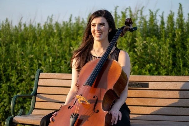 A cello instructor for in home lessons serving Glenview, Northbrook, Skokie, Morton Grove and Winnetka with long dark hair playing a cello outdoors on a park bench, with a green field and trees in the background.