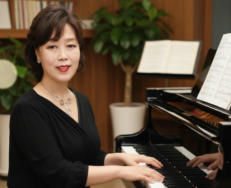 A Chicago piano teacher for piano lessons in your home in Chicago suburbs at a piano with sheet music, indoors with a plant and bookshelf in the background.