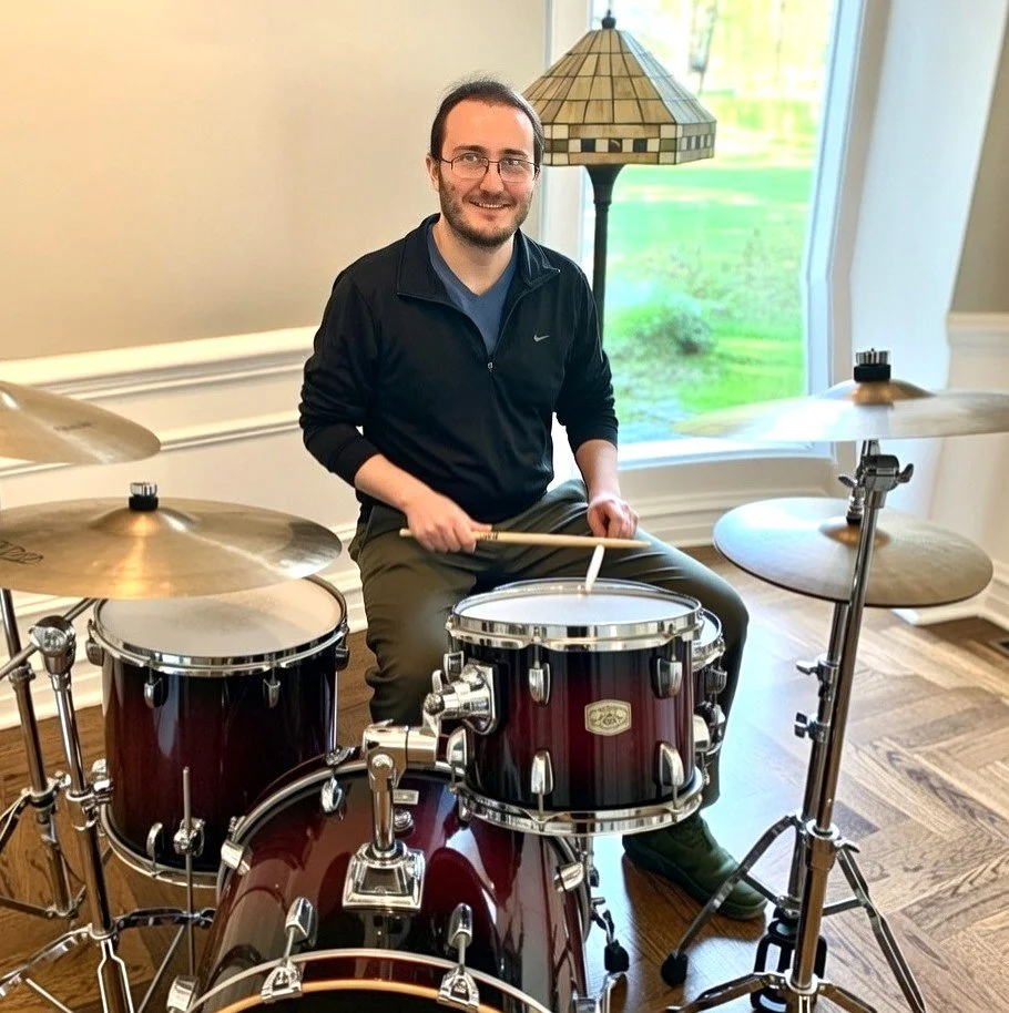 A Chicago area in-home music teacher who teaches percussion lessons and ukulele lessons sitting behind a drum set, smiling, with a window and a lamp in the background.