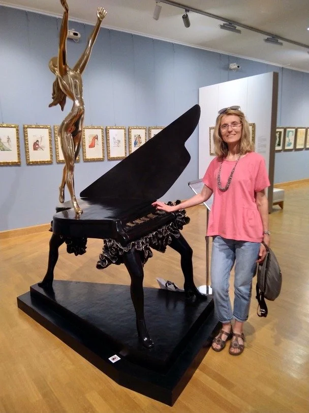 A mandolin teacher who offers mandolin lessons in your home for Chicago suburbs stands next to a piano sculpture with a woman figure on top, in an art gallery with framed artwork on the wall.