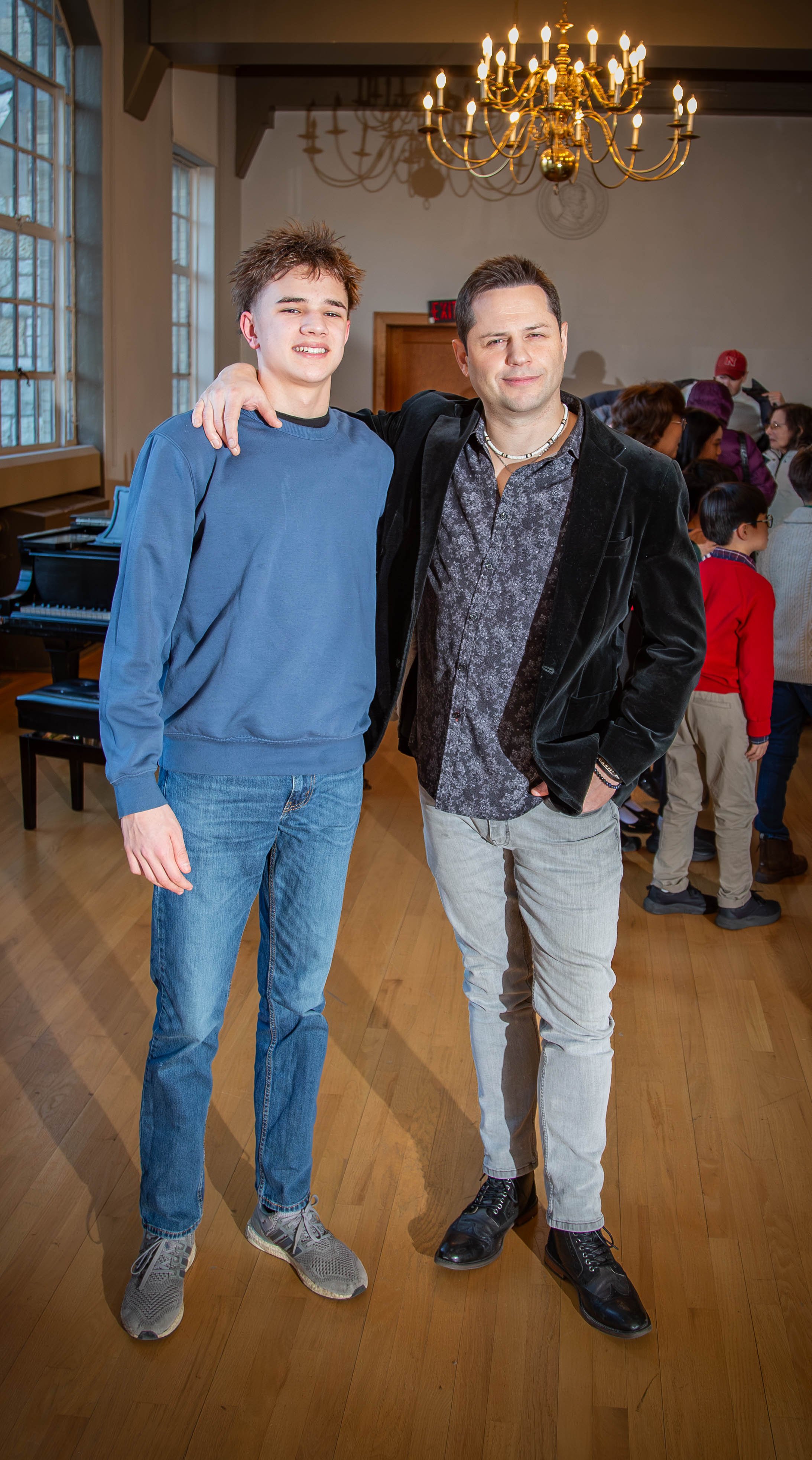 Two men standing together in a room with a chandelier and a piano, with a group of children and adults in the background.