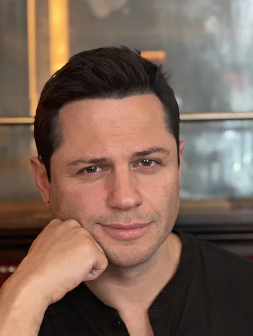 A man with dark hair styled back, resting his chin on his hand, wearing a black shirt, and smiling subtly at the camera.
