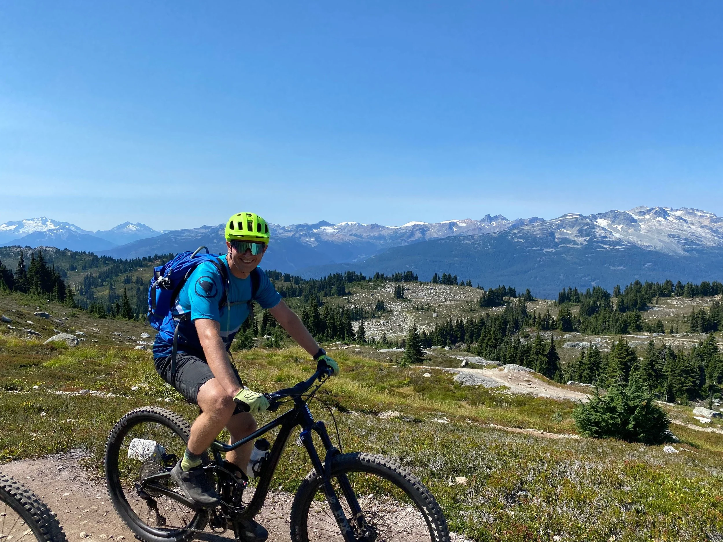A man mountain biking on a trail in a scenic mountainous area with snow-capped peaks in the background, wearing a bright yellow helmet, sunglasses, a blue shirt, and a backpack.