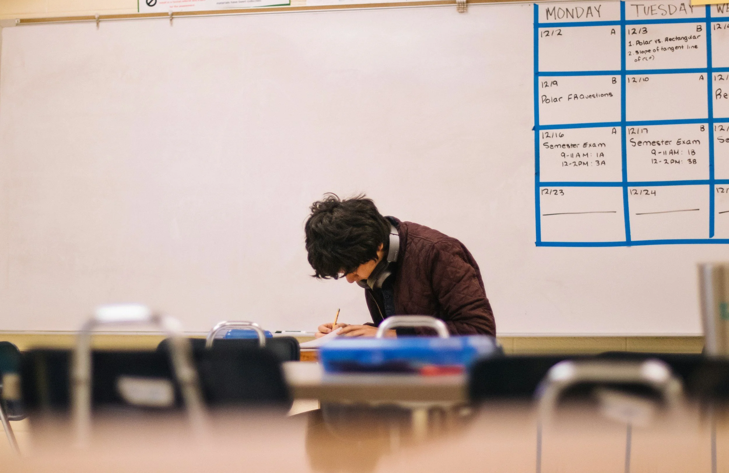 A student with headphones working at a classroom desk, with a whiteboard and a schedule on the wall in the background.
