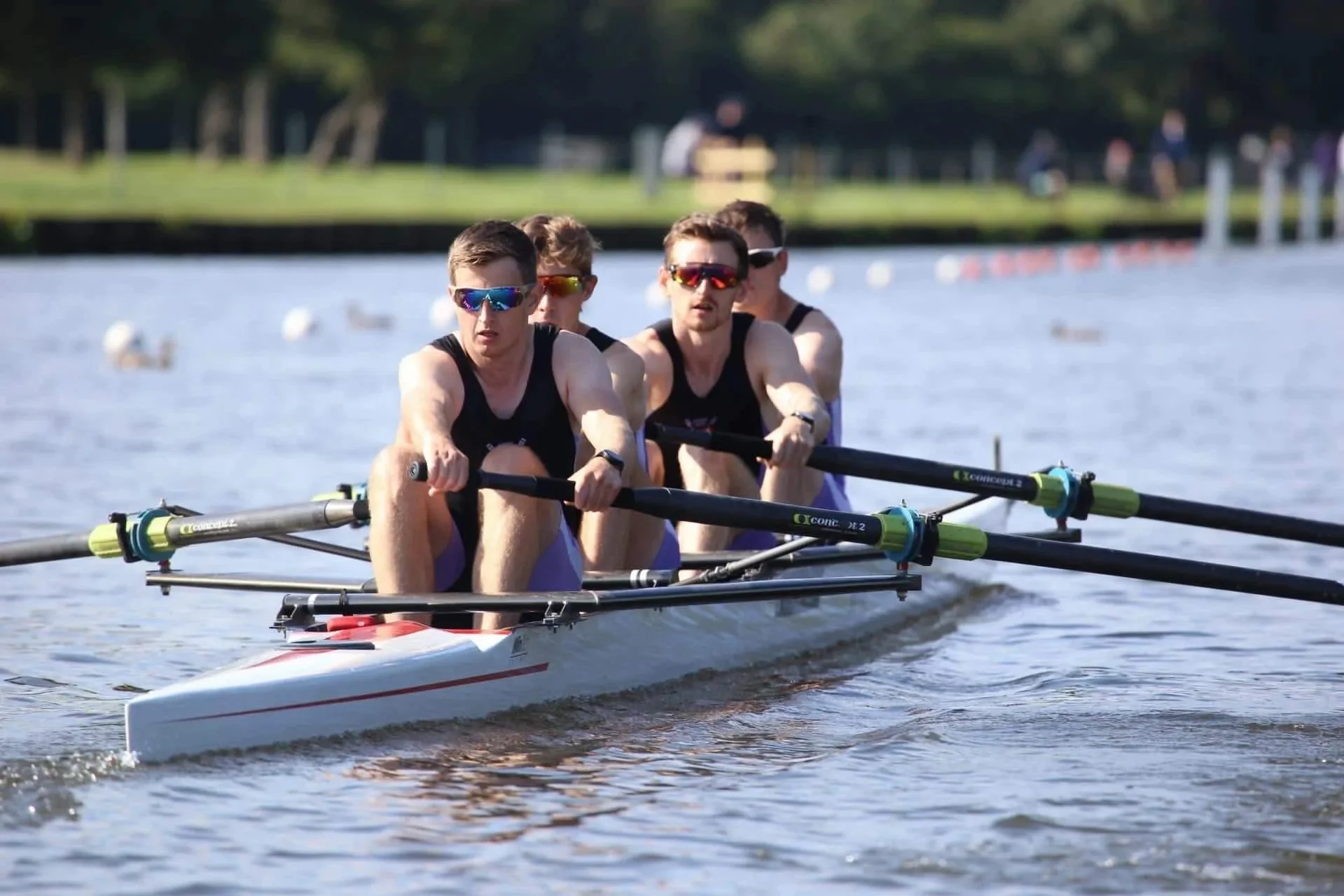 A team of four rowers in a racing shell, wearing black sleeveless shirts and sunglasses, rowing on a body of water.