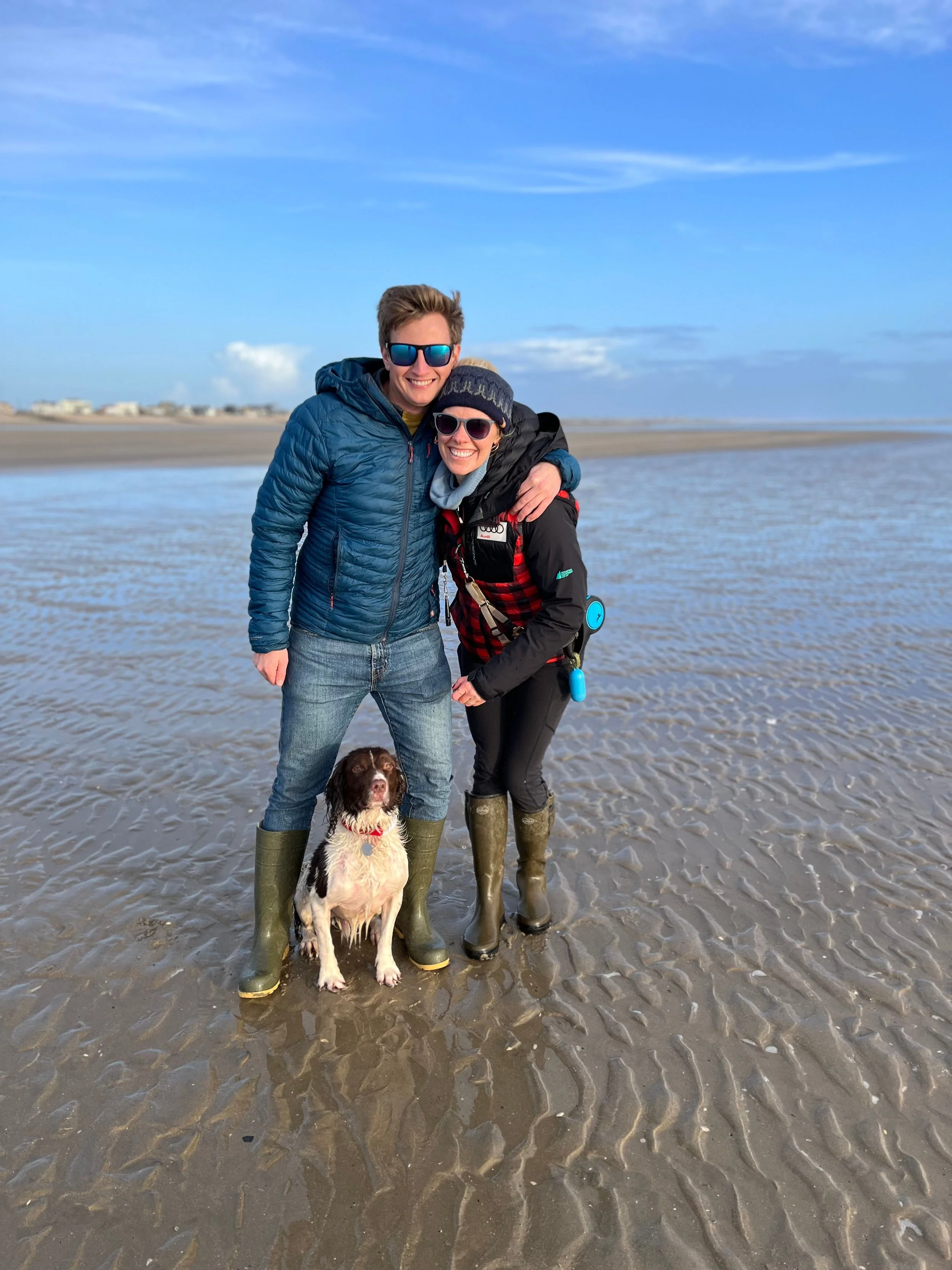 A smiling couple with a dog stands on a wet sandy beach with tide pools. The man has short brown hair, wears a blue jacket, jeans, and green rubber boots. The woman has dark hair, wears a black and red jacket, a knit hat, sunglasses, and green rubber