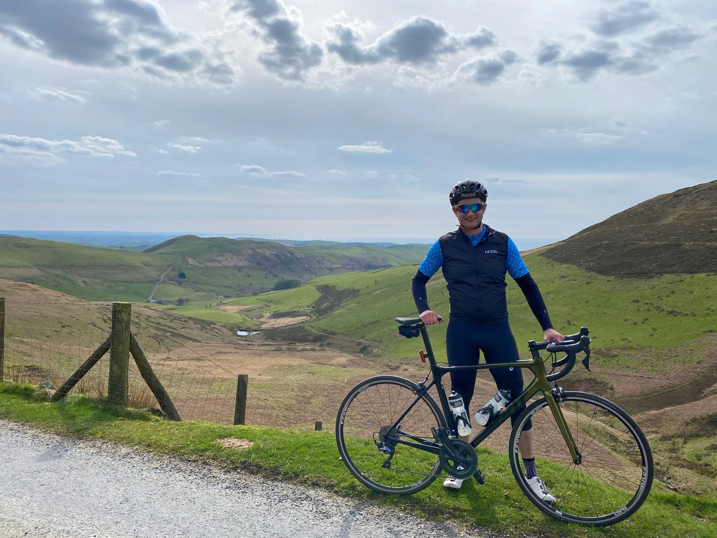 A cyclist standing next to a black bike on a grassy roadside overlooking rolling green hills under a partly cloudy sky.