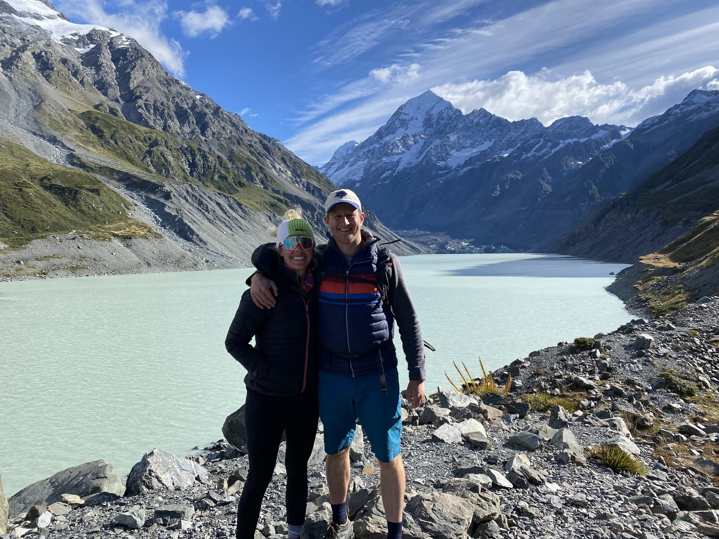 A smiling couple stands together in front of a mountain lake surrounded by snow-covered peaks, rocky terrain, and blue sky with wispy clouds.