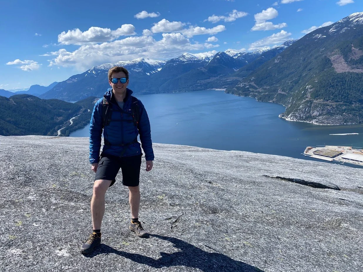 A person standing on a large rocky surface with a mountain and water body in the background, snow-capped mountains, and partly cloudy sky.