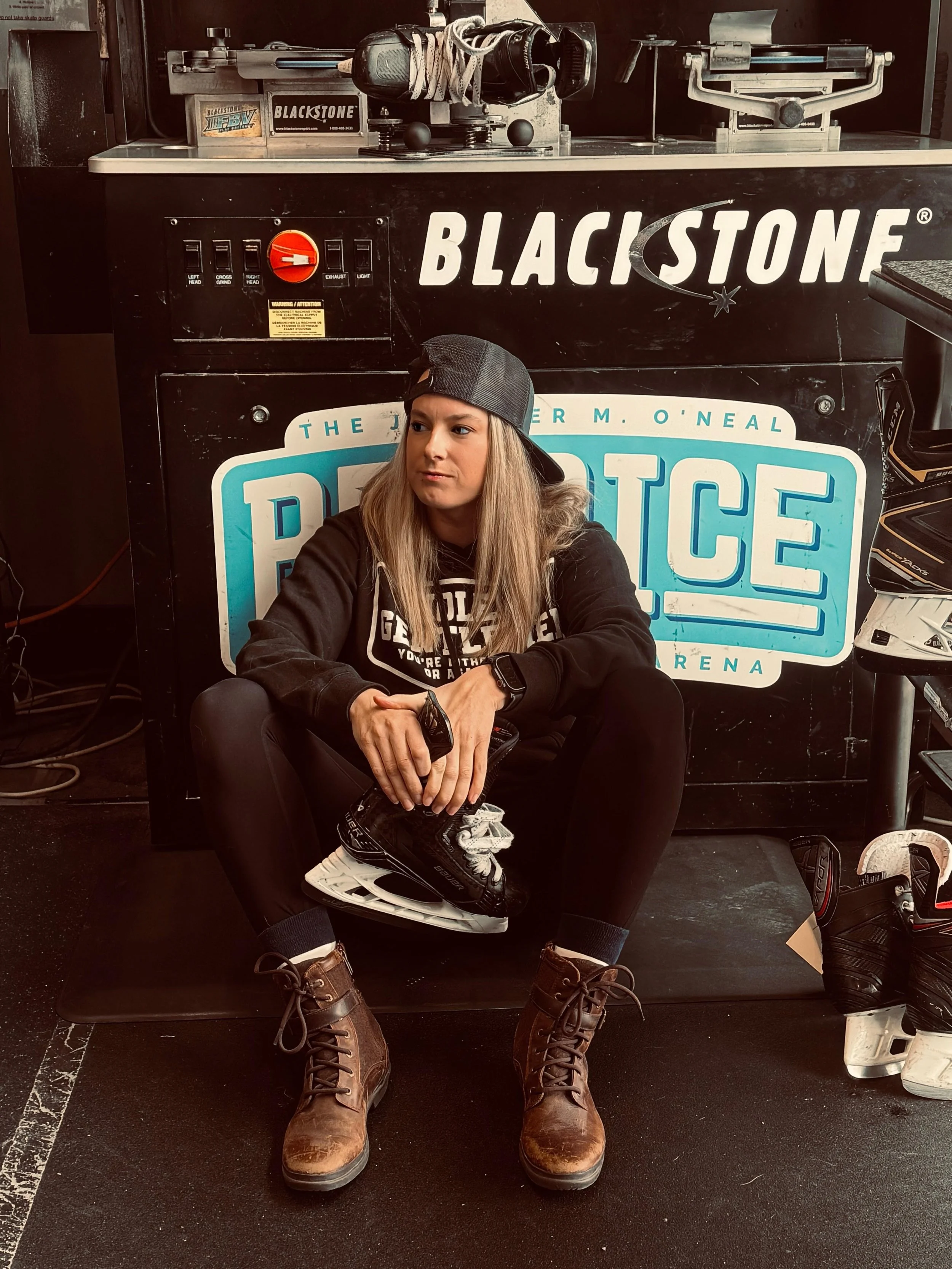 A woman sitting on the floor in a hockey skate store, holding a hockey skate, wearing a black cap backwards, black hoodie, black leggings, and brown boots, with hockey equipment and gear around her.