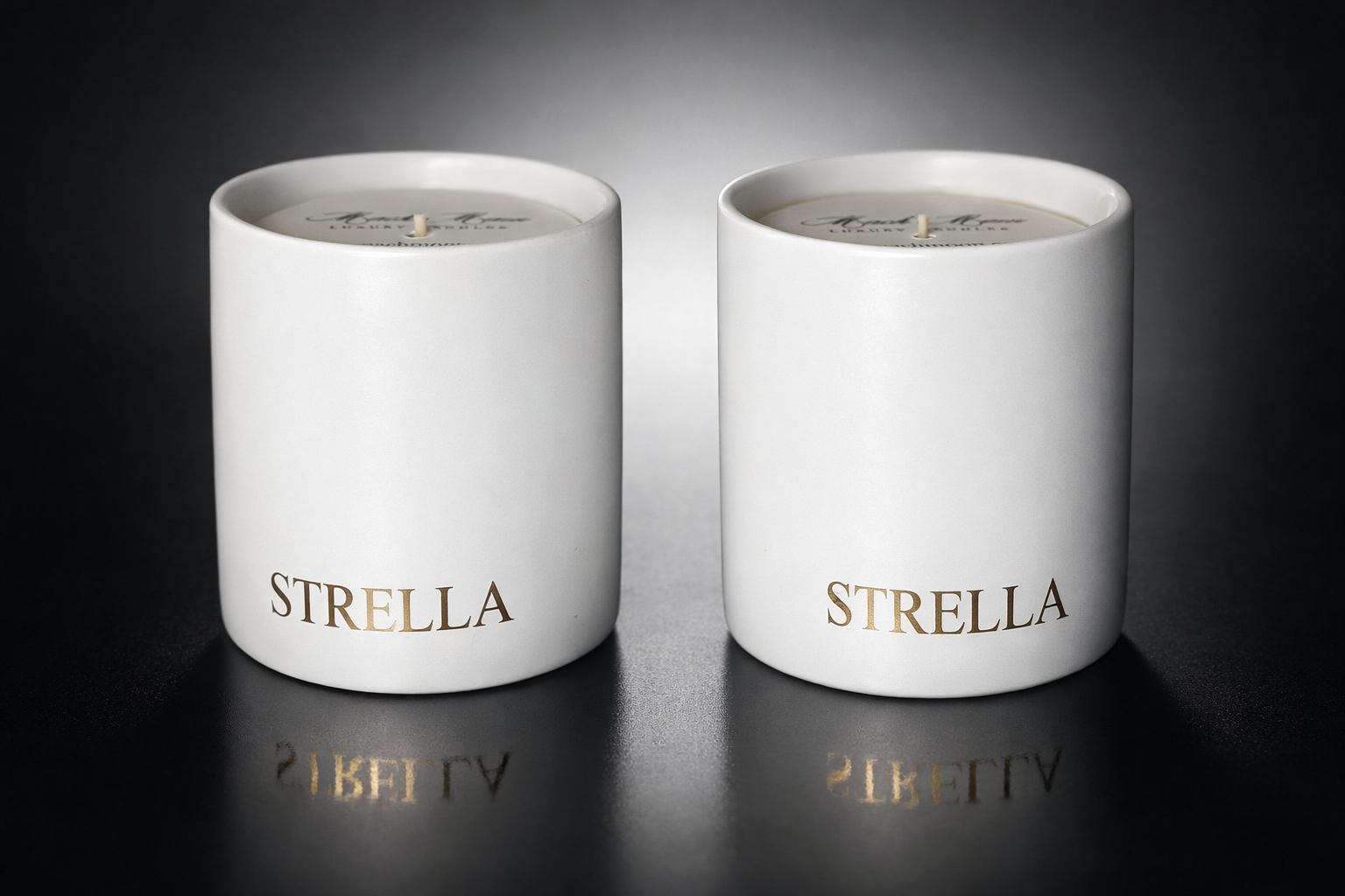 Two white candles in cylindrical holders labeled 'Strella' in gold lettering, placed on a reflective black surface with a gradient background.