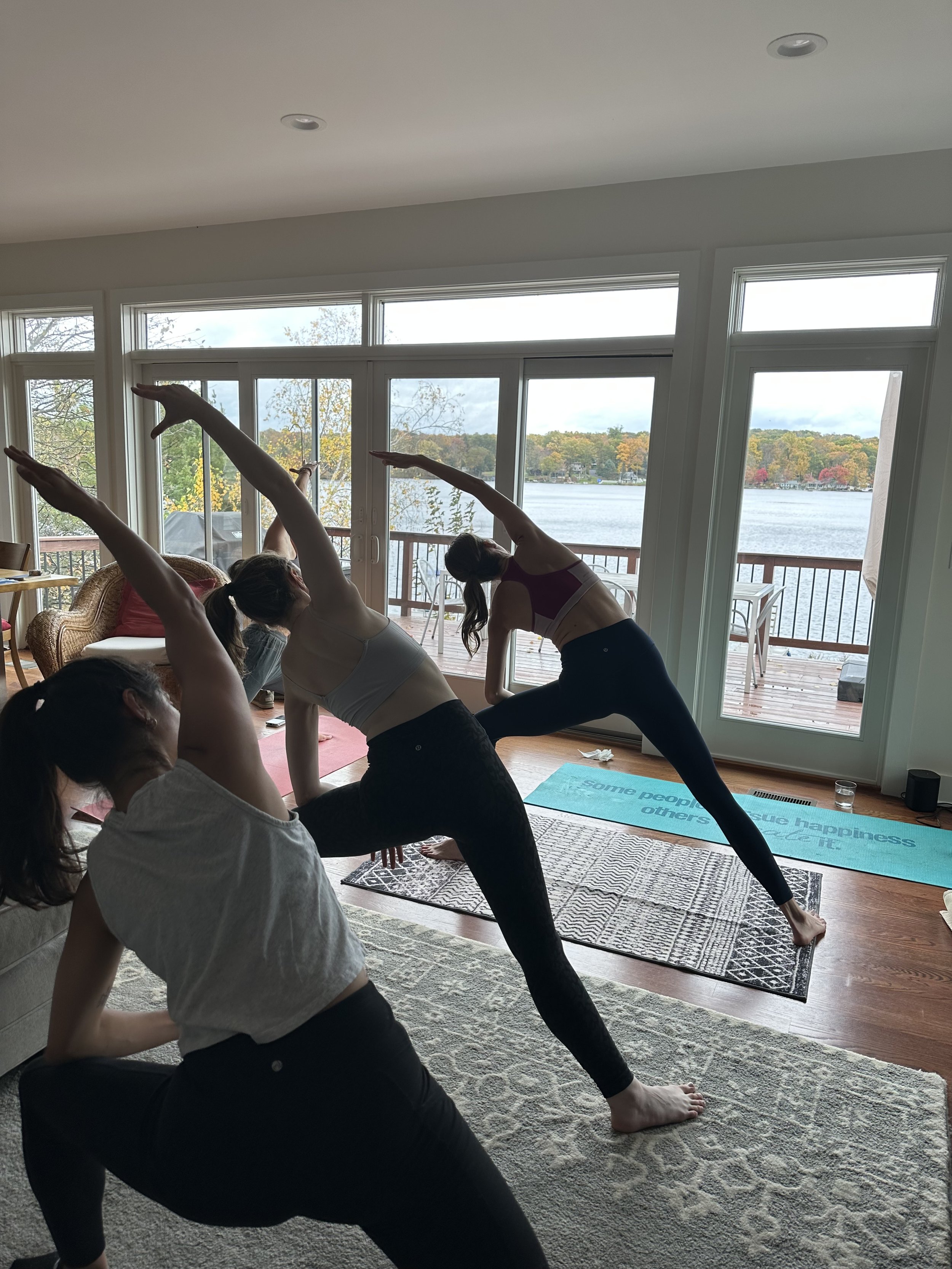 Four women practicing yoga indoors near large windows overlooking a lake, with trees and colorful foliage in the background.