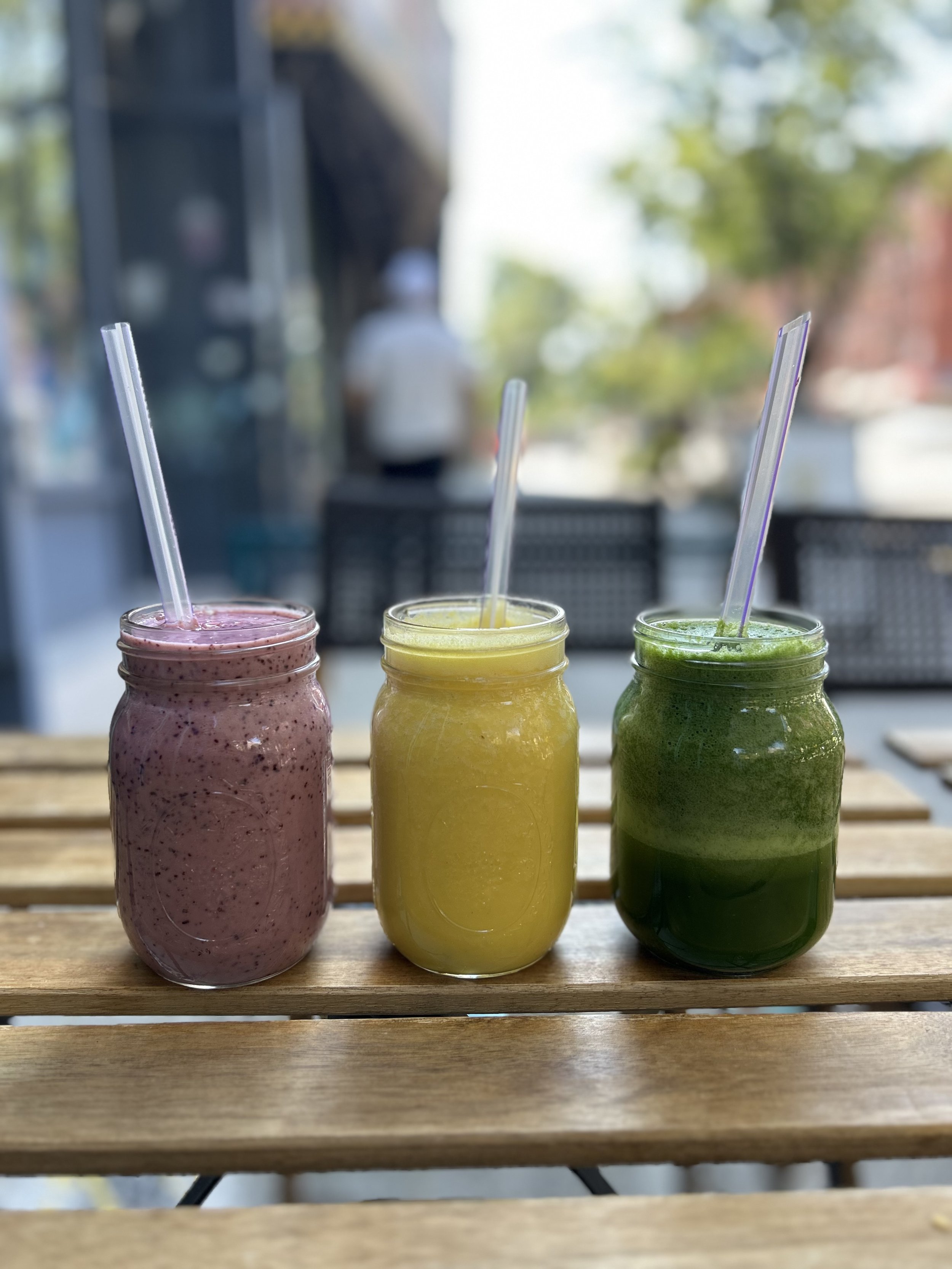 Three mason jars filled with smoothies in pink, yellow, and green colors, each with a clear straw, sitting on a wooden table outdoors.