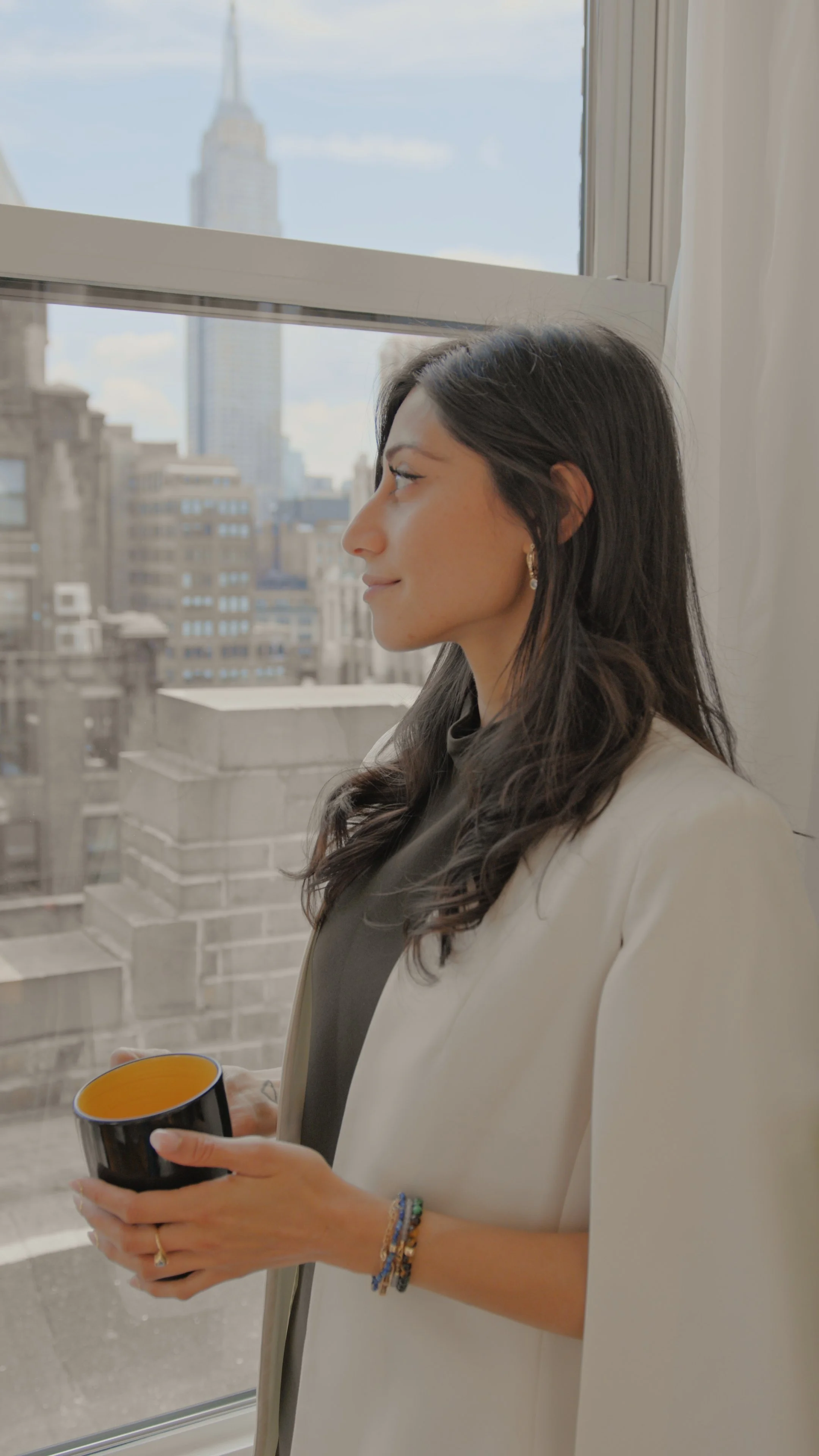 A woman with dark, wavy hair and earrings standing by a window, holding a black mug with an orange interior, with a city skyline in the background.