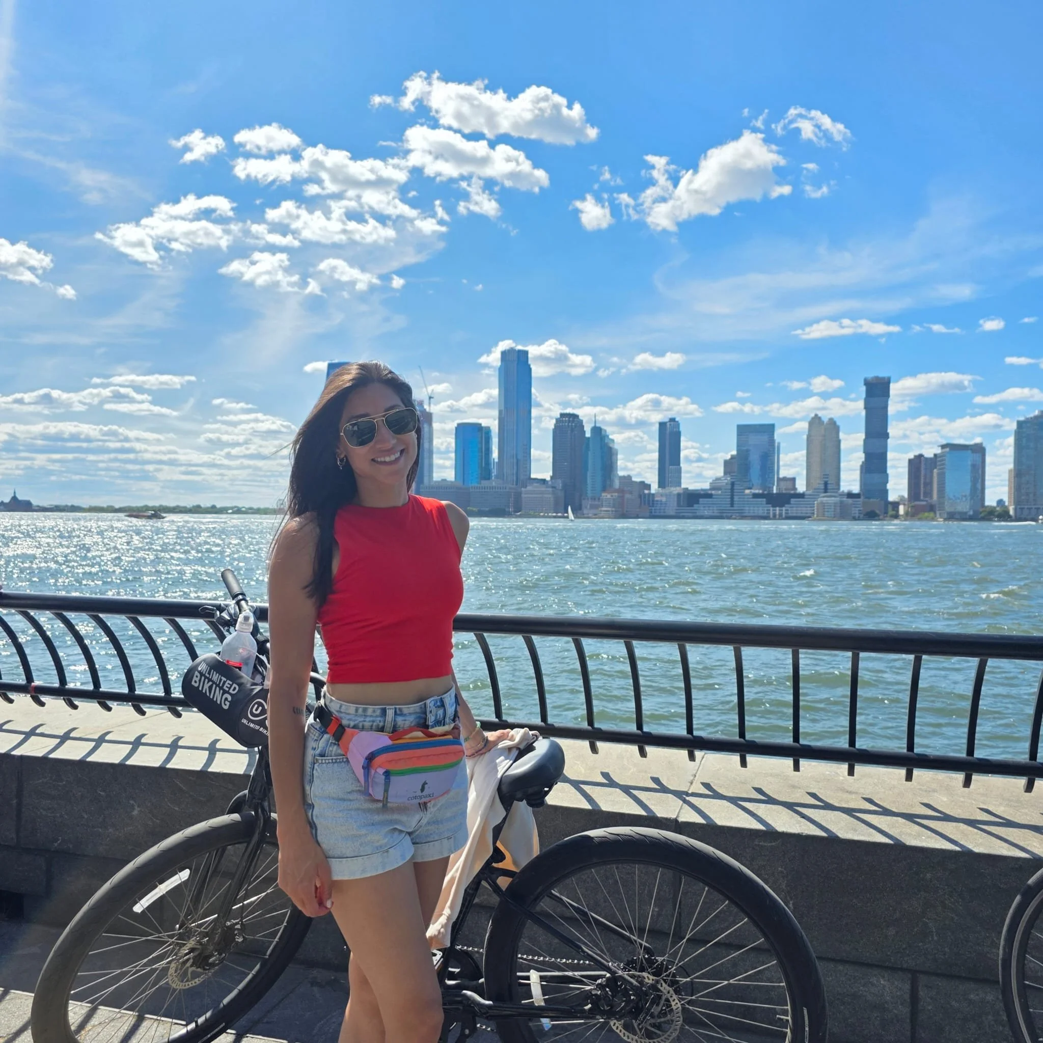 A young woman in a red sleeveless top and denim shorts, wearing sunglasses and a rainbow fanny pack, standing beside a bicycle along a waterfront with a city skyline in the background on a sunny day.