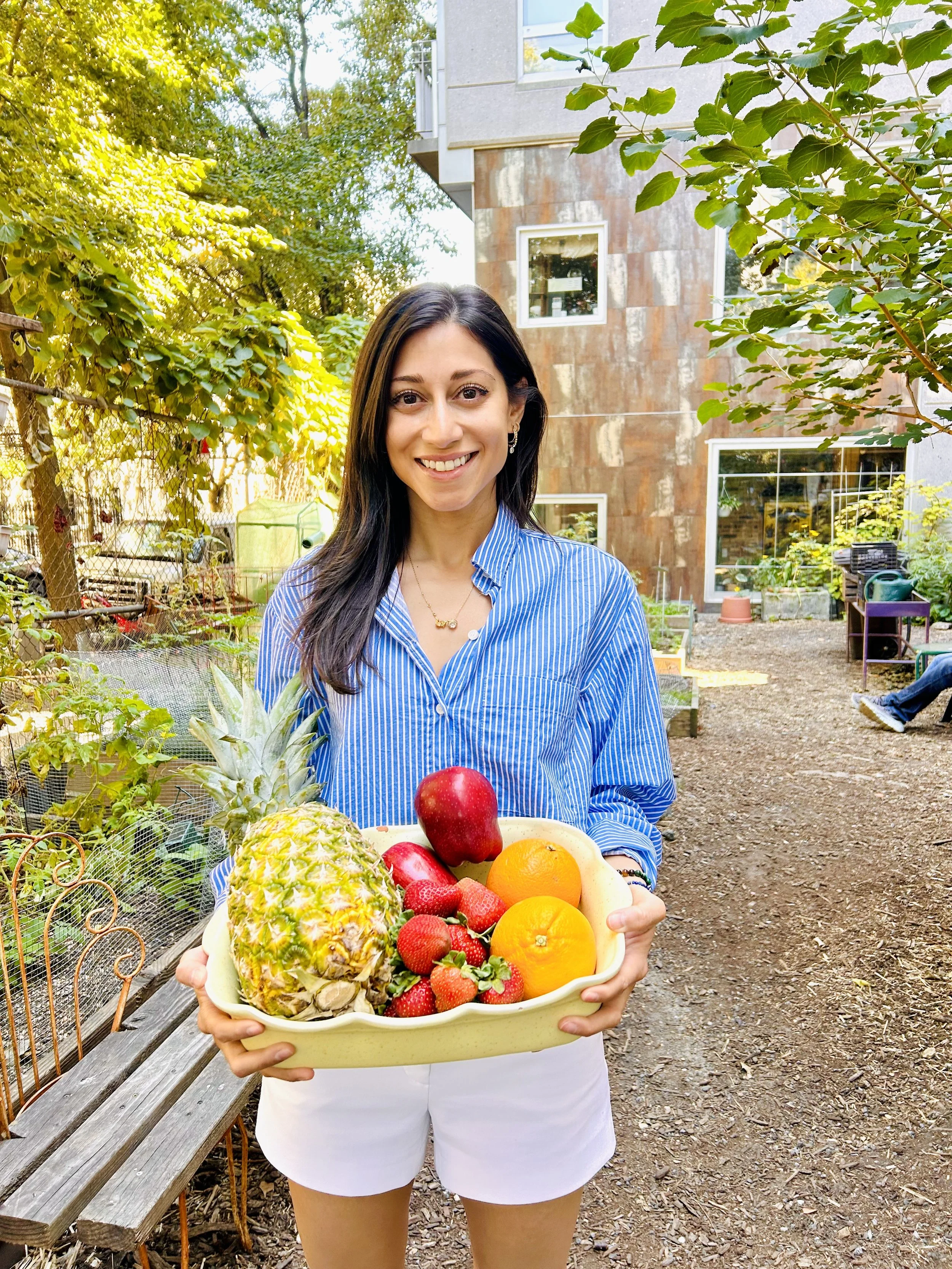A woman with long dark hair and a blue striped shirt smiling while holding a tray of fresh tropical fruits, including pineapple, apples, oranges, and strawberries, in an outdoor garden setting with trees, plants, and a modern building in the background.