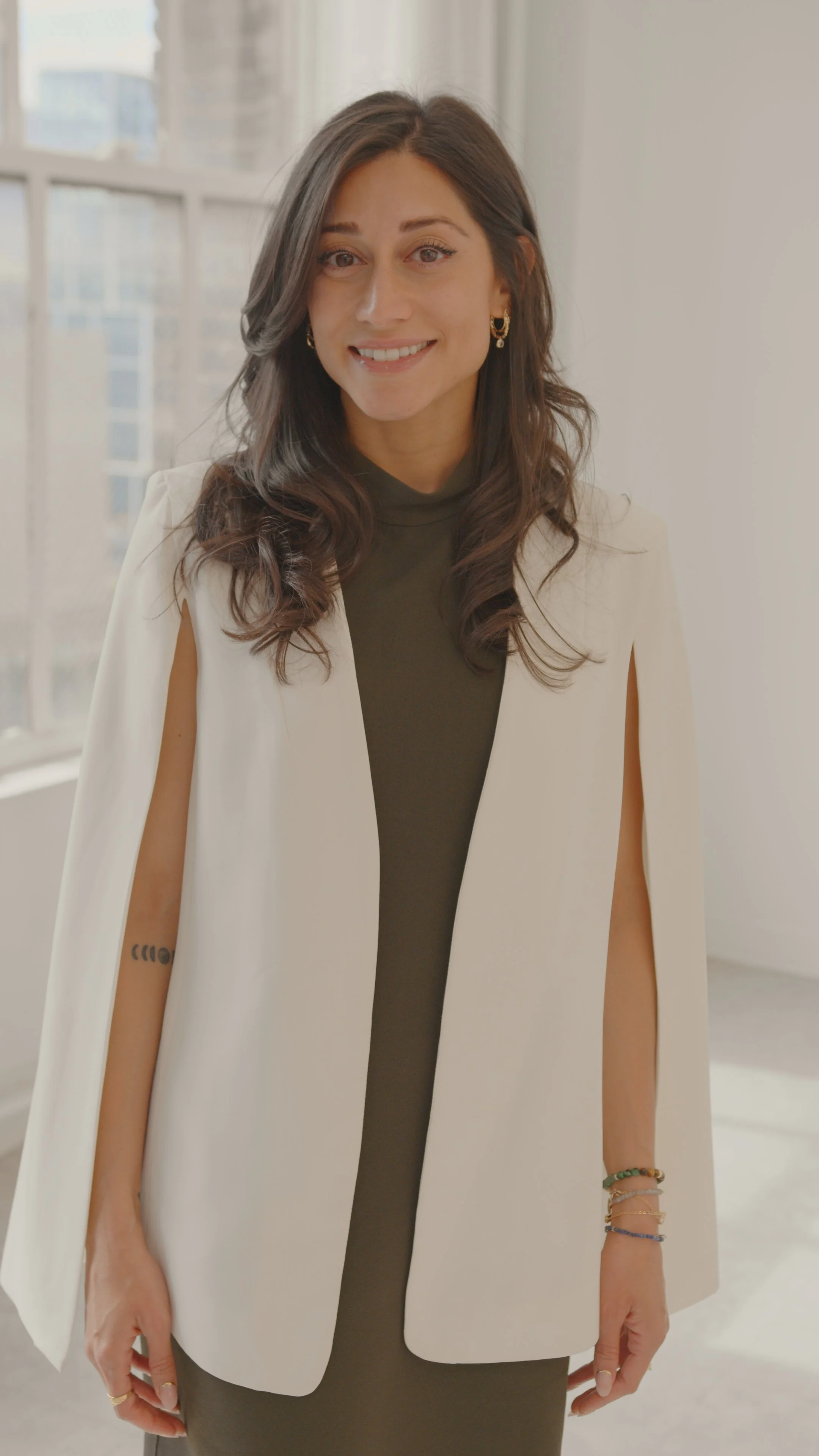 A woman with dark brown hair, wearing a black top and a white sleeveless blazer, standing in front of a window in a bright room, smiling at the camera.