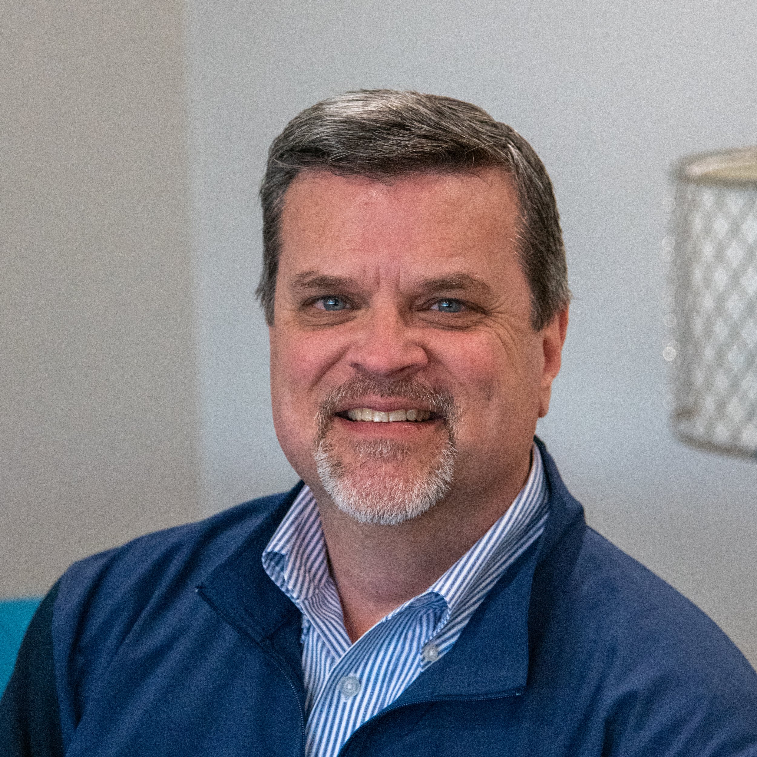 A middle-aged man with gray hair and a beard, smiling, wearing a blue jacket and a striped shirt, sitting indoors.