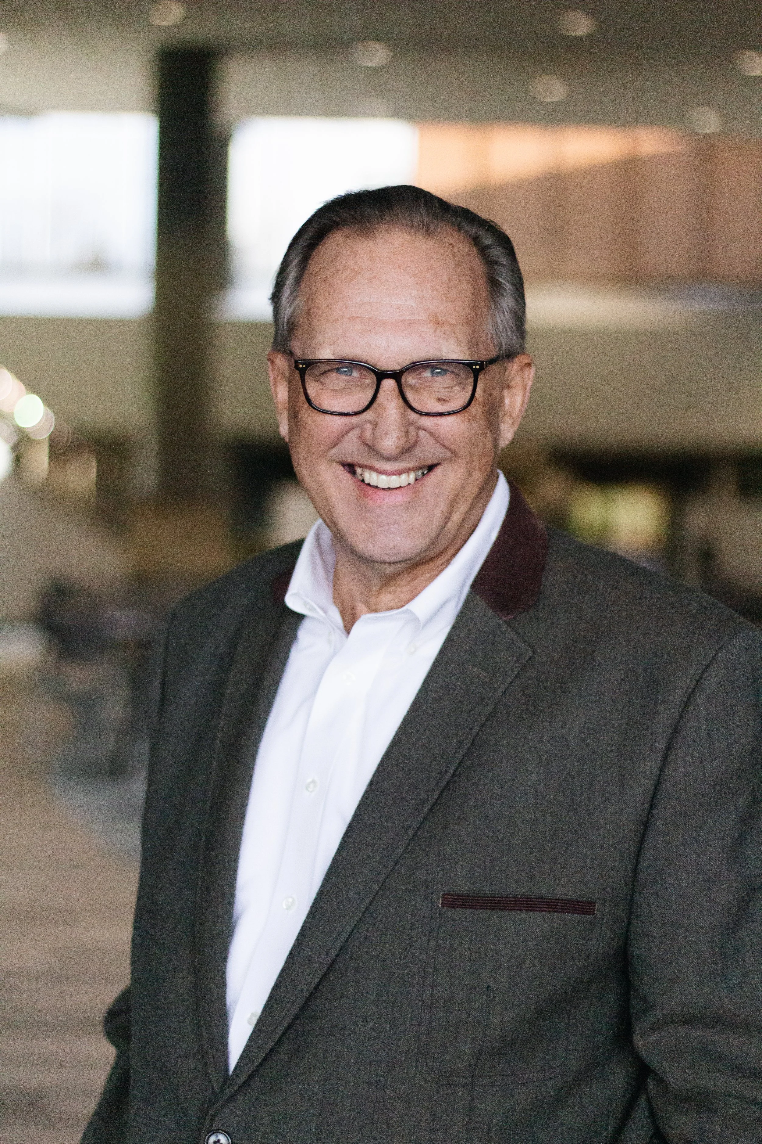 A smiling middle-aged man wearing glasses, a white shirt, and a dark gray blazer, standing indoors in a modern building.