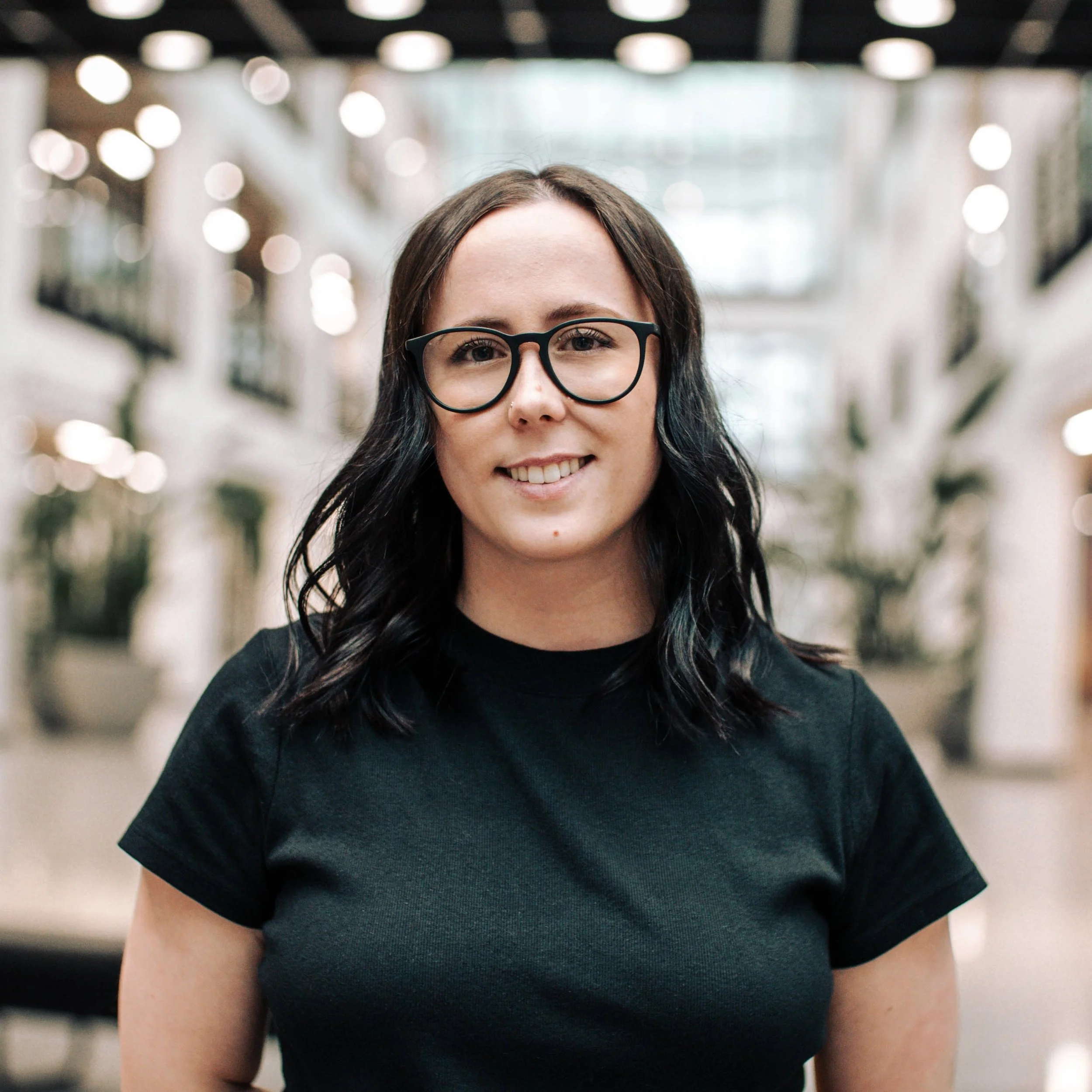 A young woman with dark wavy hair wearing glasses and a black t-shirt, smiling in an indoor space with blurred lights and architecture in the background.